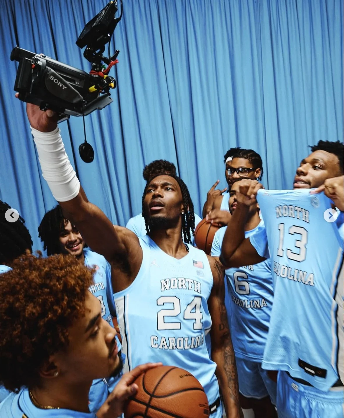 A group of North Carolina basketball players taking a selfie after a game or practice, with a player holding a camera and other players holding basketballs and making gestures.