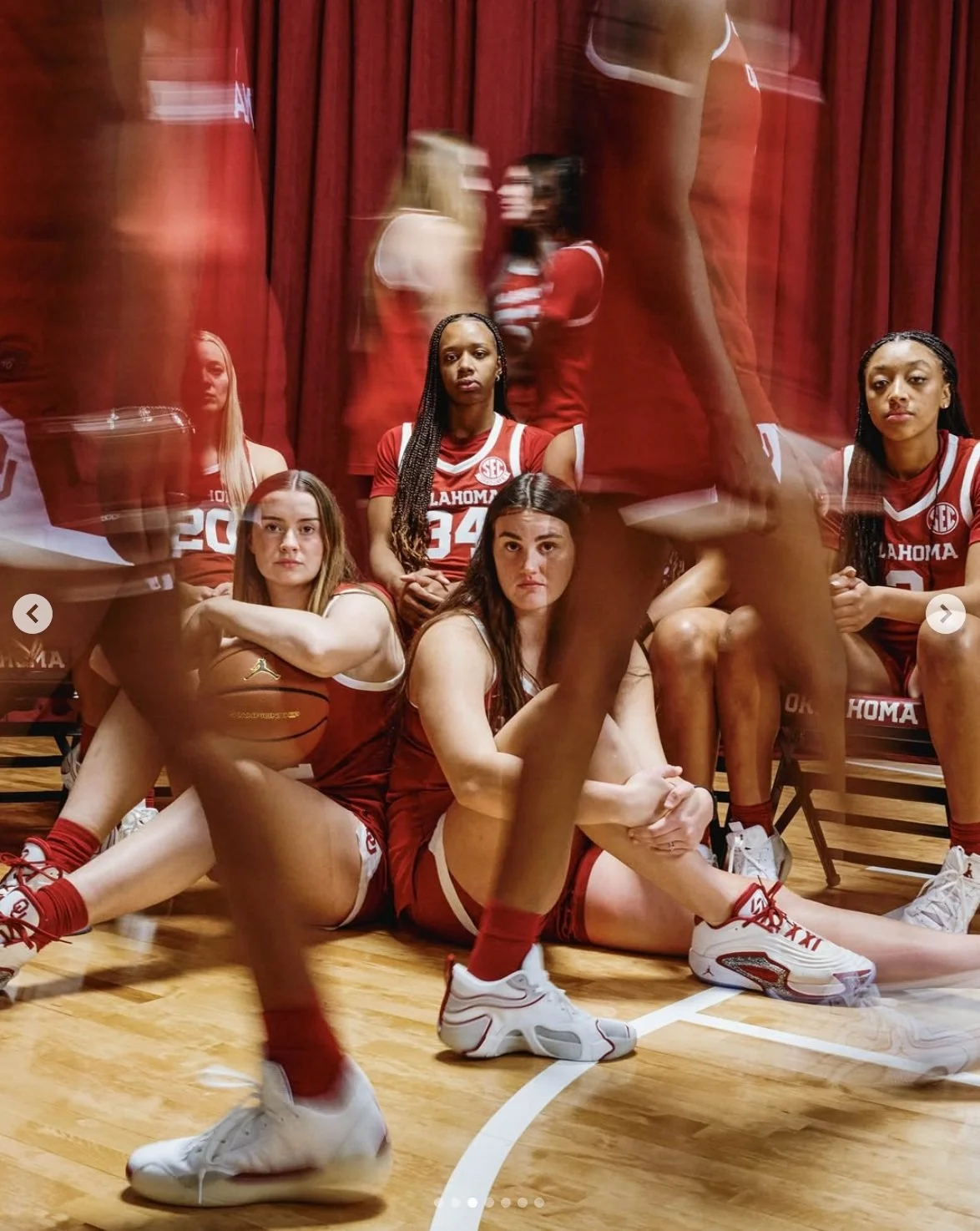 Group of female basketball players in red Oklahoma jerseys sitting and standing in a gym near a red curtain, with motion blur of a player's legs in the foreground.