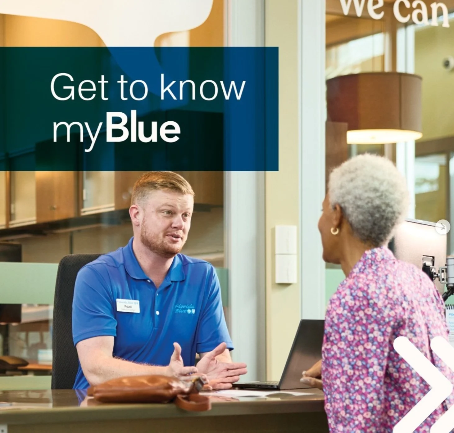 A man in a blue Florida Blue shirt and an elderly woman are talking at a reception desk in an office or healthcare setting. There is a sign that says 'Get to know my Blue'.