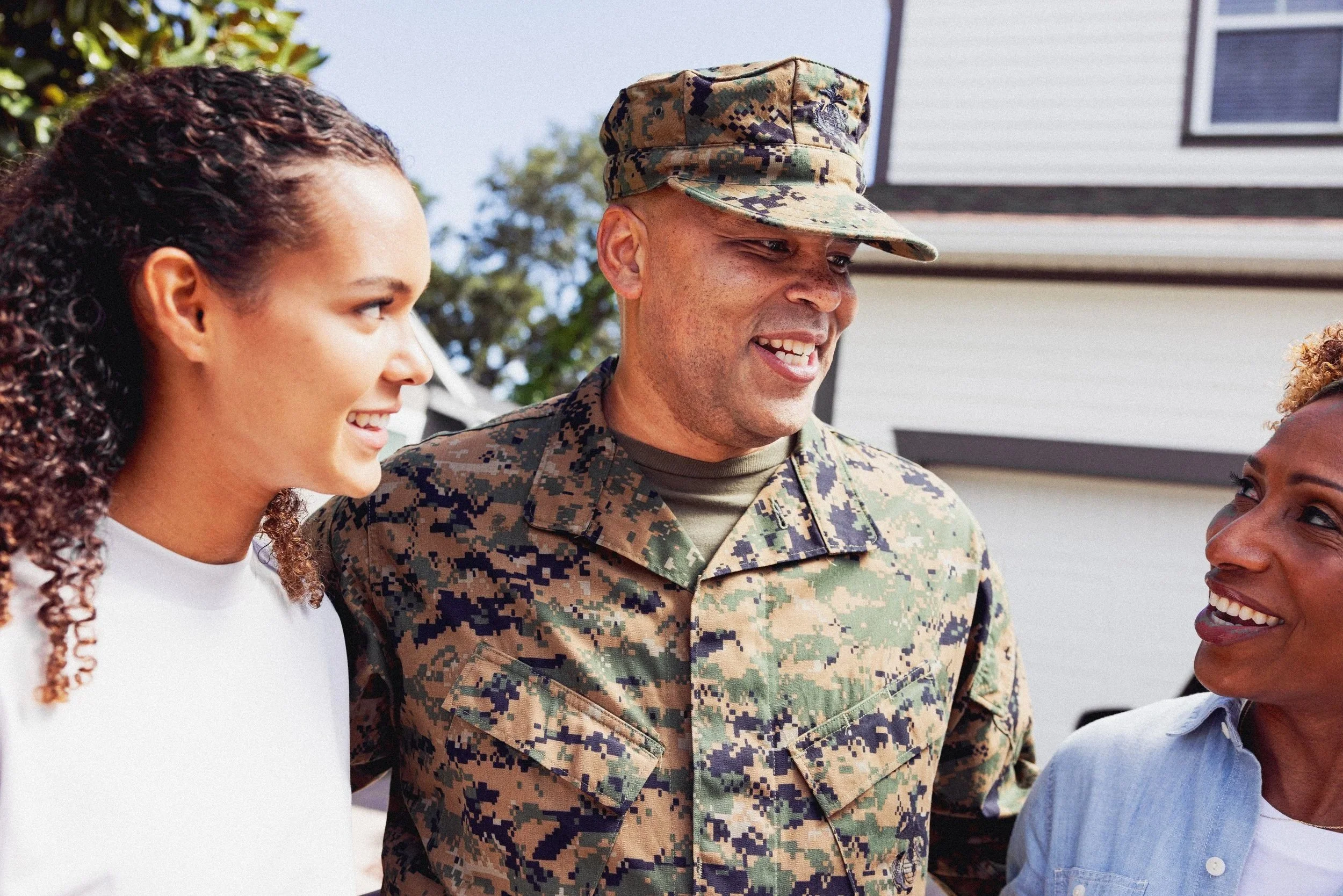 A male soldier in a camouflage uniform talking with his wife and daughter outside their home.