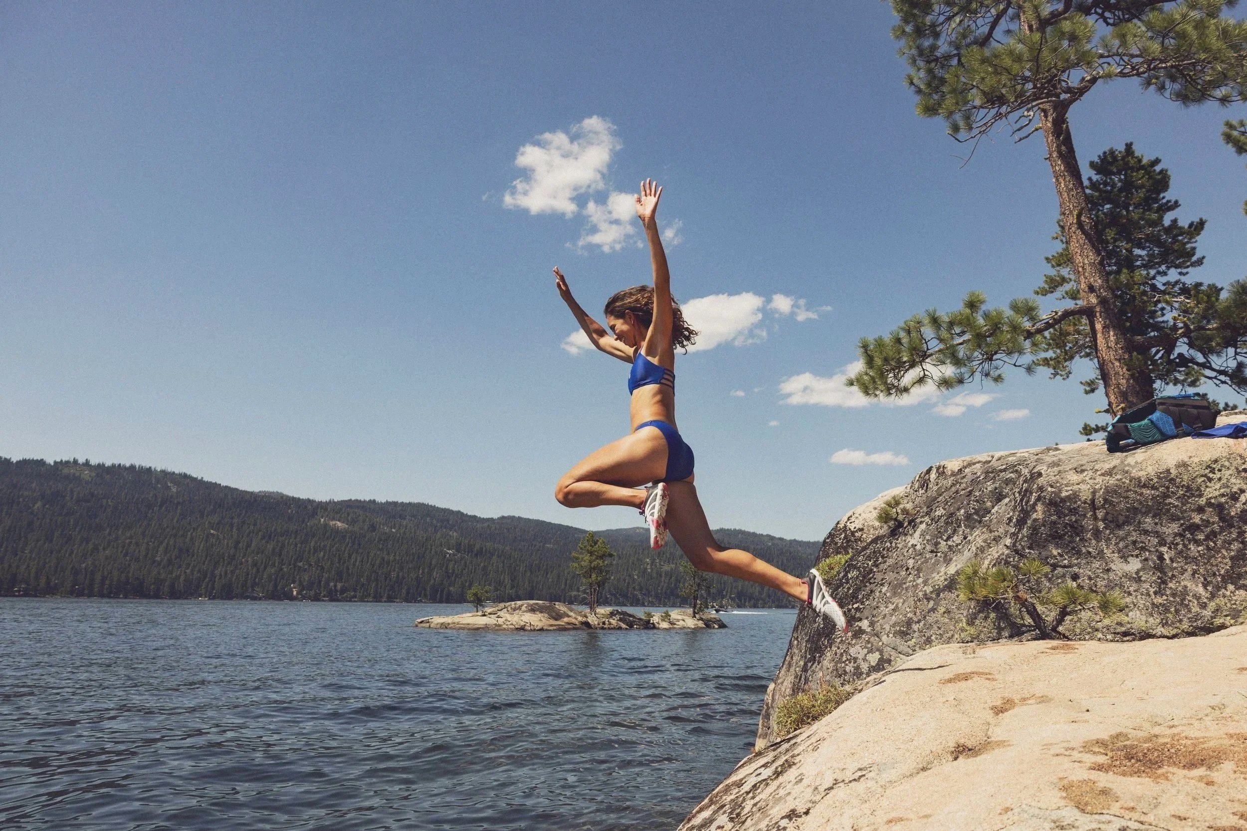 A woman in a blue sports bra and shorts jumping in the air on a large rock by a lake, with pine trees and mountain hills in the background under a blue sky with a few clouds.
