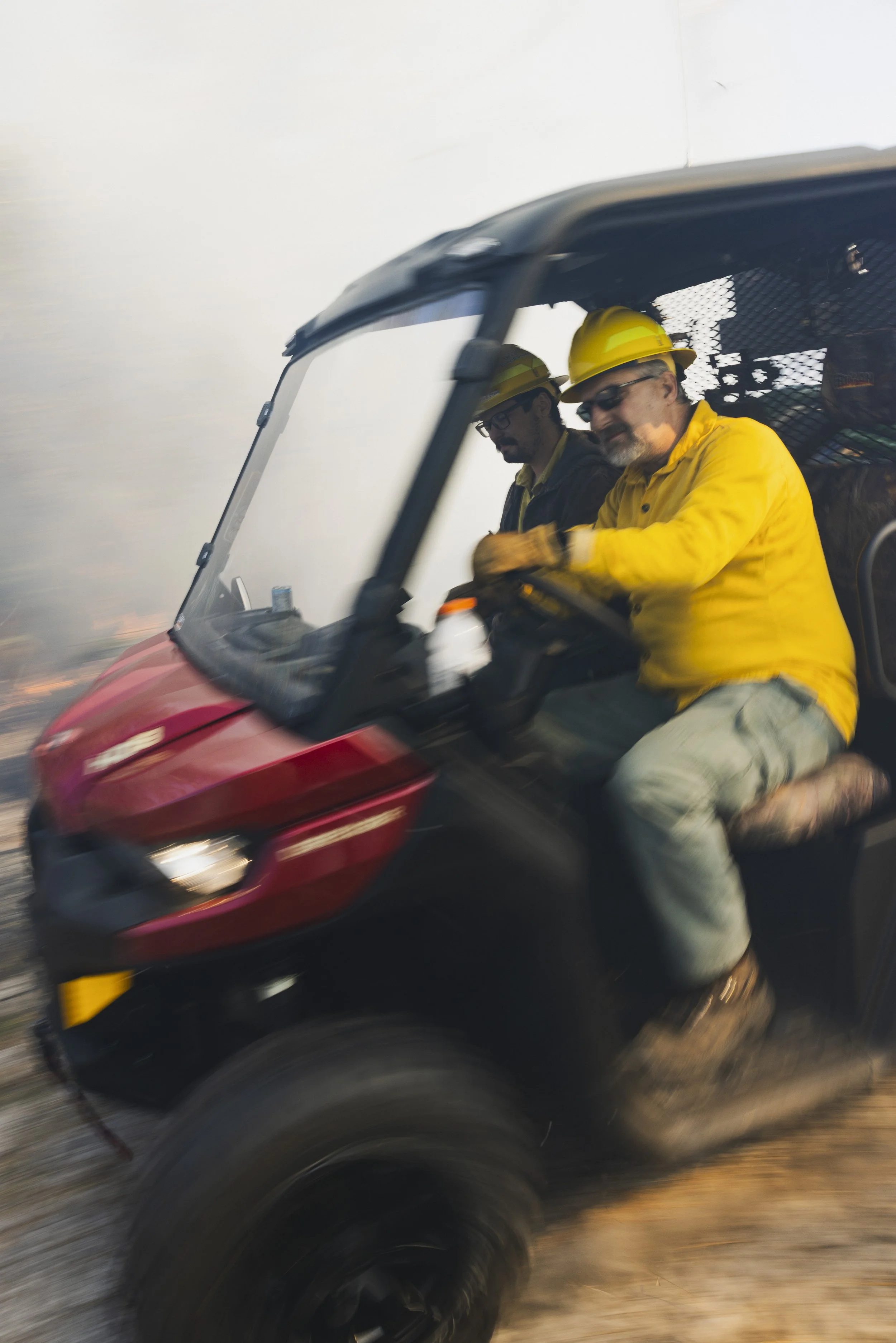 Two workers wearing yellow safety helmets and yellow safety jackets riding a red utility vehicle through a smoky environment.