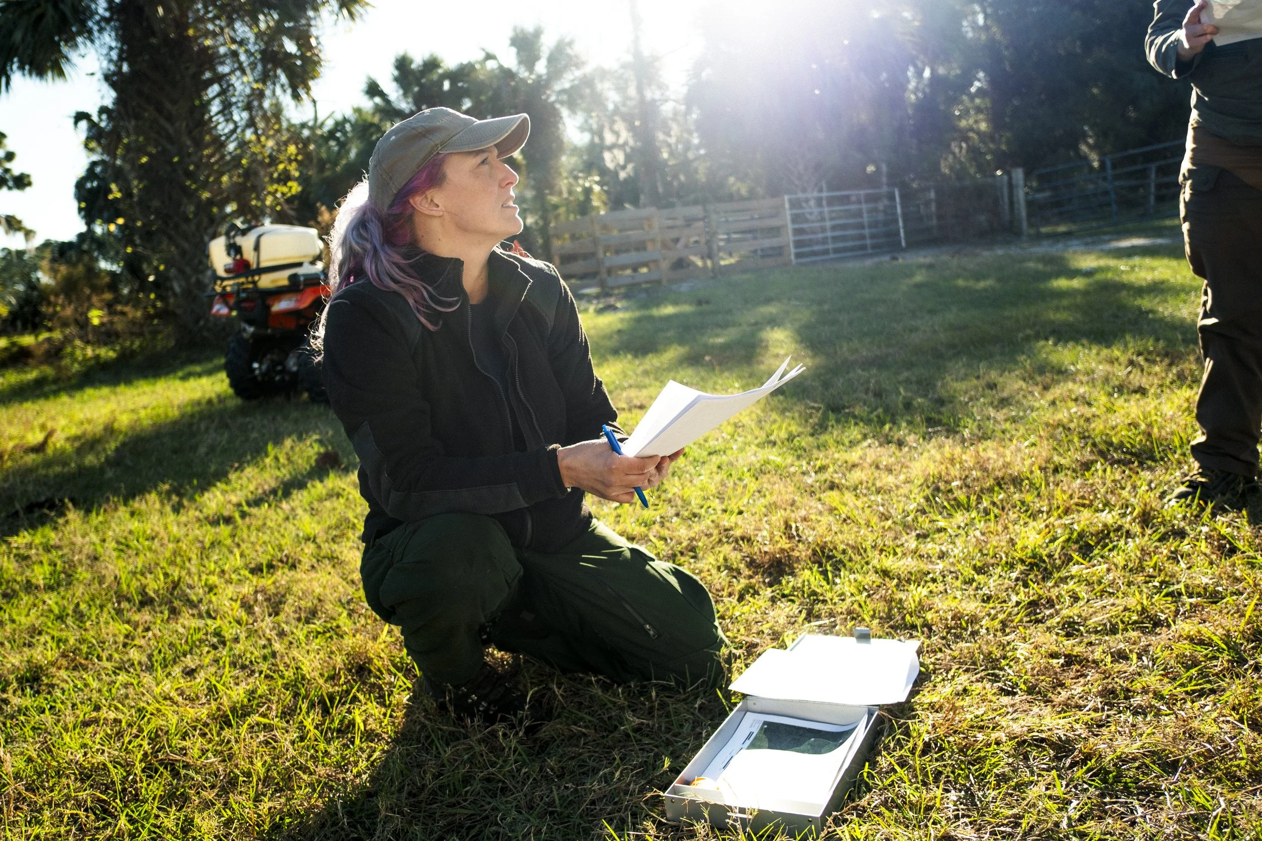 A woman in outdoor gear, kneeling on grass, holding papers and a pen, with a box on the ground, outdoors with sunlight and trees in the background.