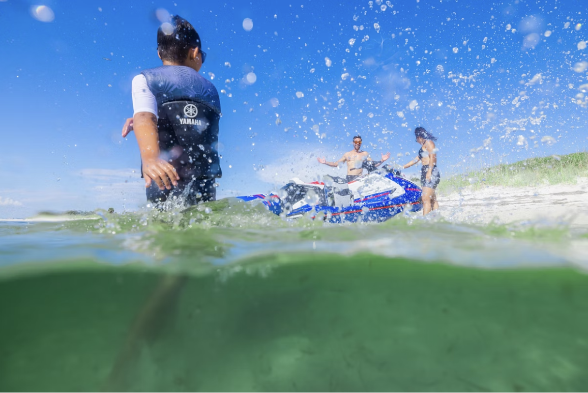People at the beach with a jet ski in the water, splashing, enjoying a sunny day.