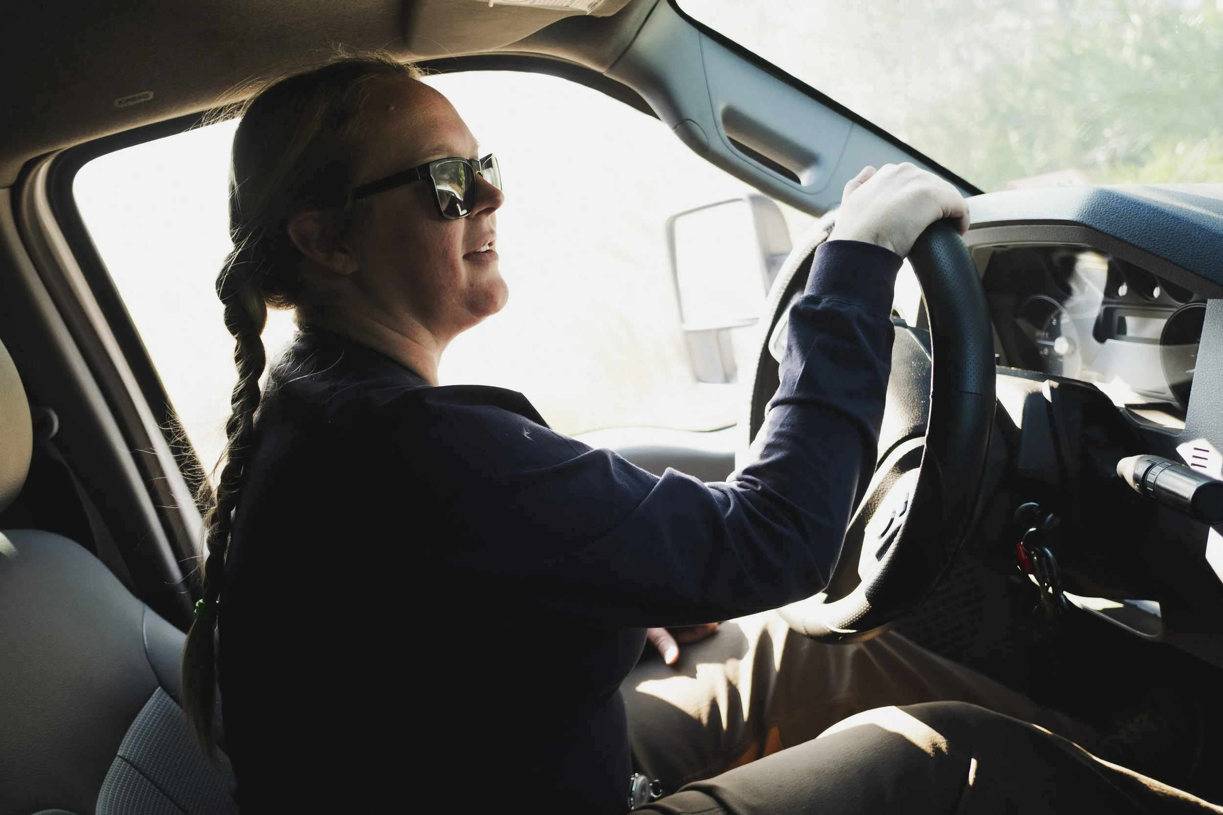 Woman with braided hair wearing sunglasses sitting in the driver's seat of a vehicle, holding the steering wheel, with sunlight coming through the window.