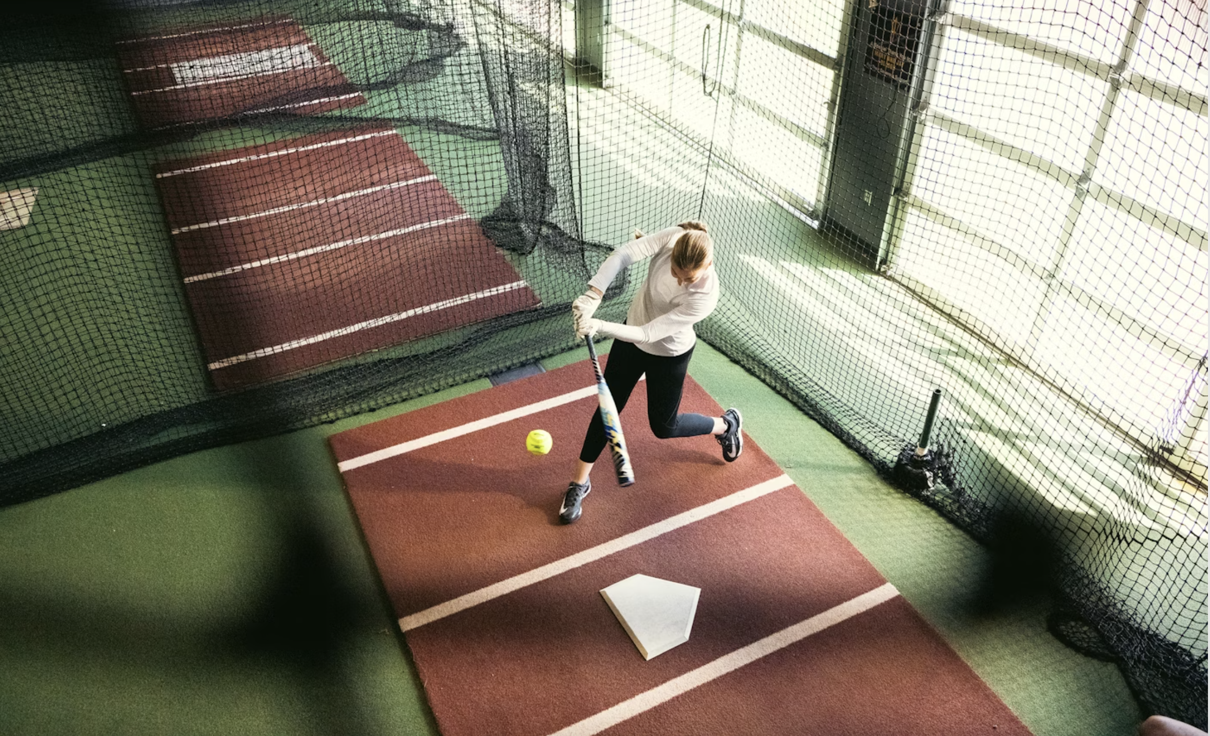 Woman practicing softball batting in indoor training facility.