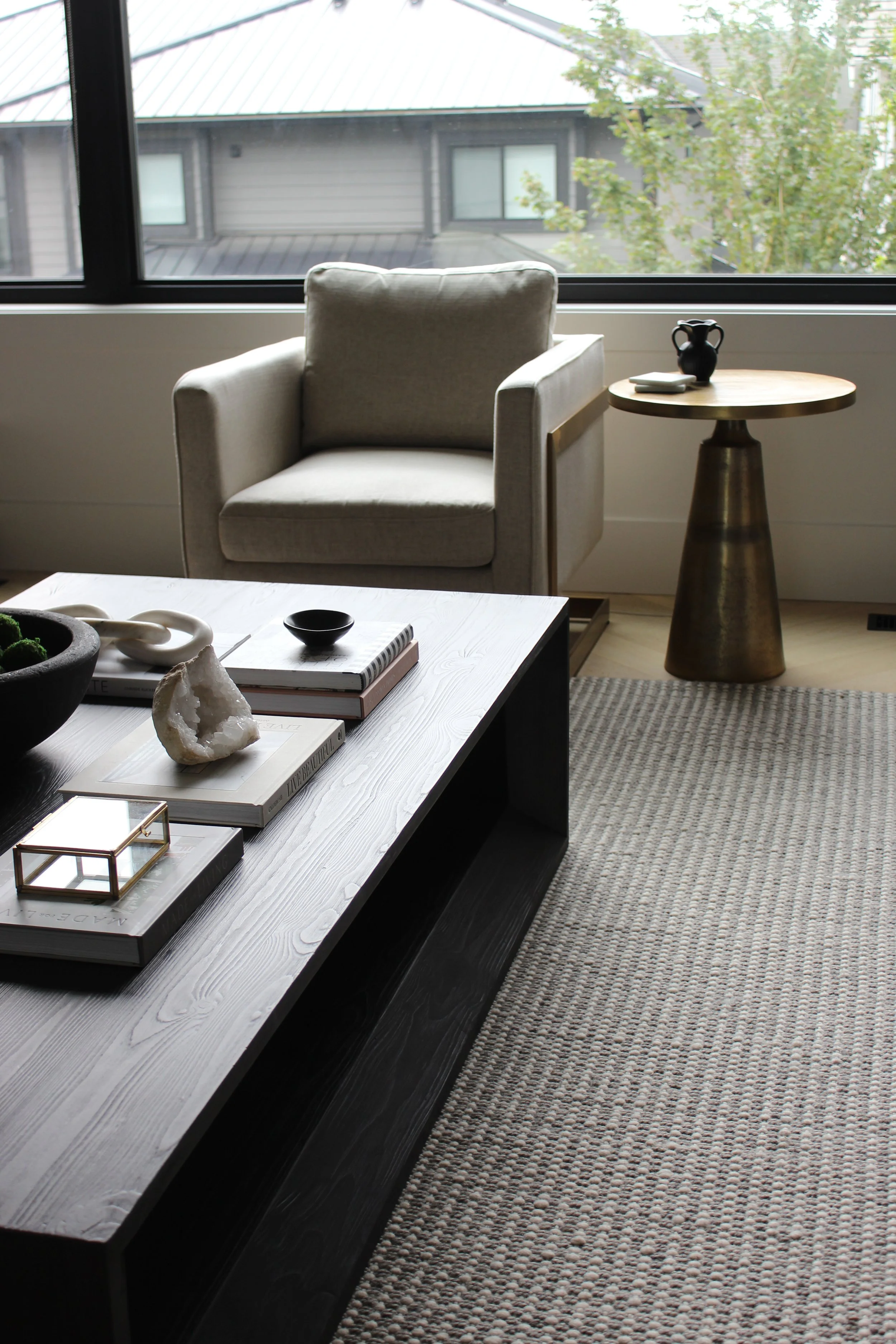 Living room with a beige armchair, a wooden side table with a black and white vase, a black coffee table with books, decorative objects, and a textured rug, set against large windows showing a neighboring house and trees outside.