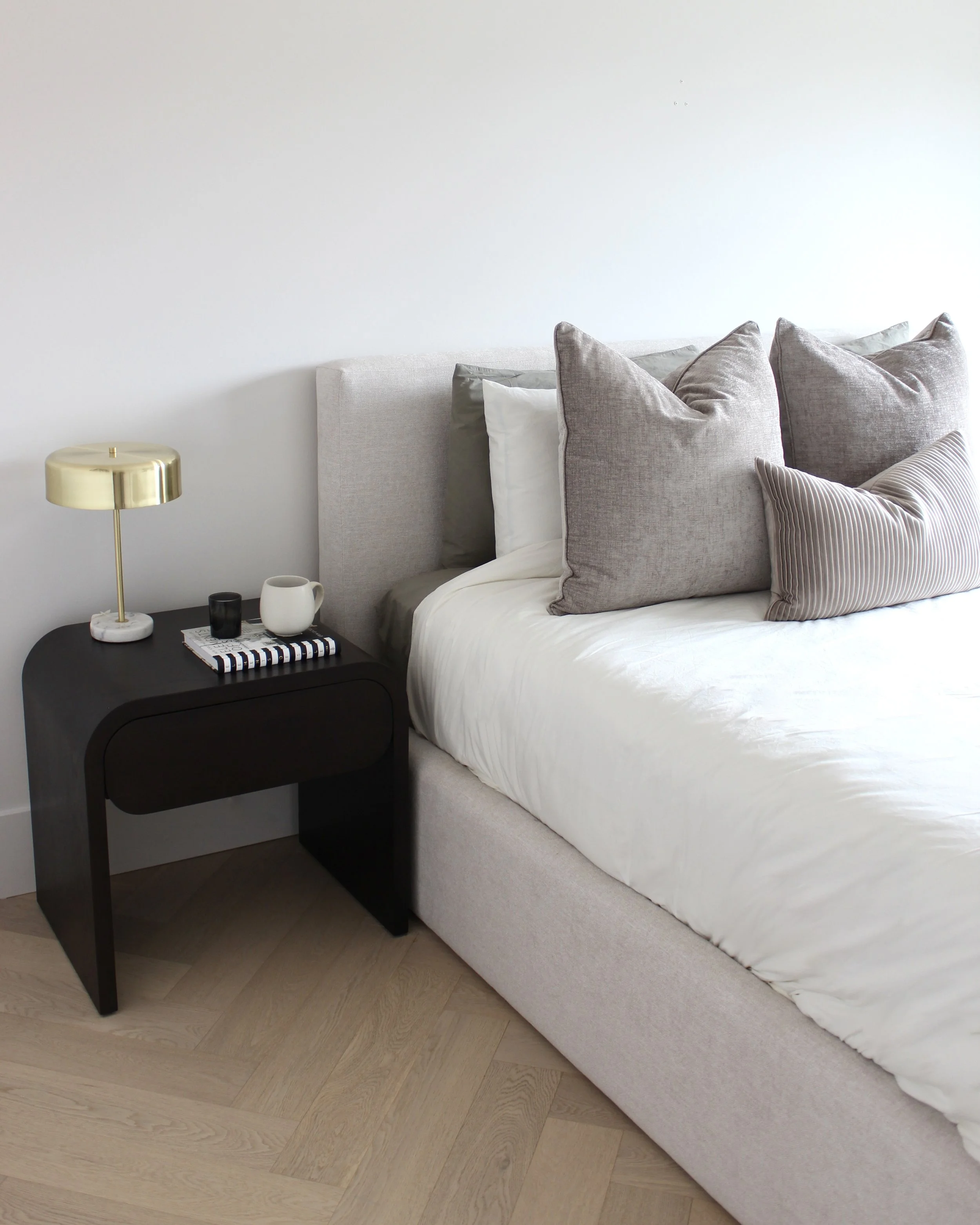 A bedroom with a bed, nightstand, and lamp. The bed has a light gray upholstered headboard, white bedding, and several gray and striped pillows. The black nightstand holds a gold-colored lamp, a black cup, a white mug, and a notebook.