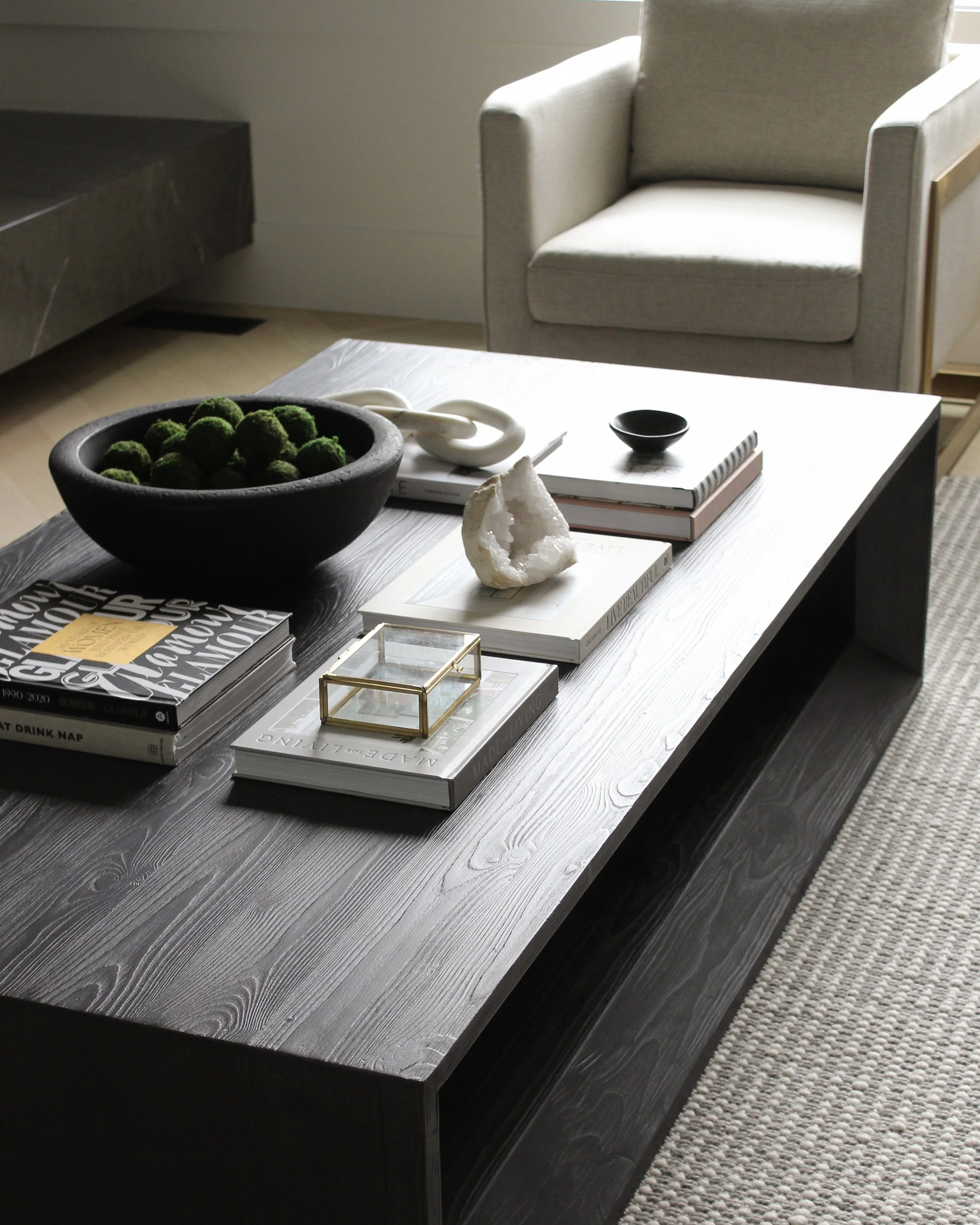 Living room with a black coffee table holding books, a black bowl of green moss balls, a white geode, a small black dish, and a glass box, near a beige armchair and a marble-topped surface.