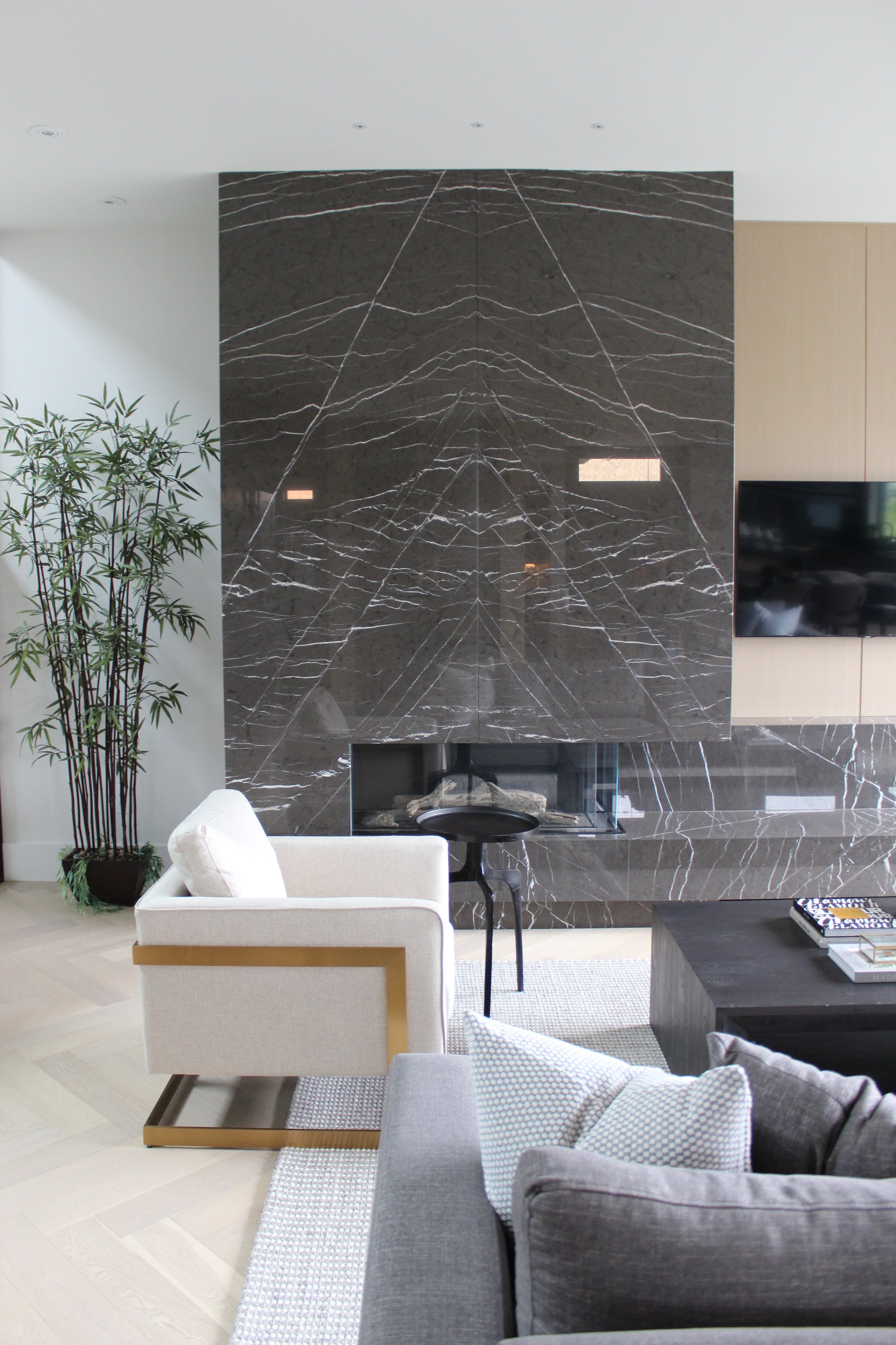 Living room with a black marble fireplace, a beige chair with gold trim, a dark gray sofa, a black side table, a flat-screen TV, and a potted plant on light wooden flooring.