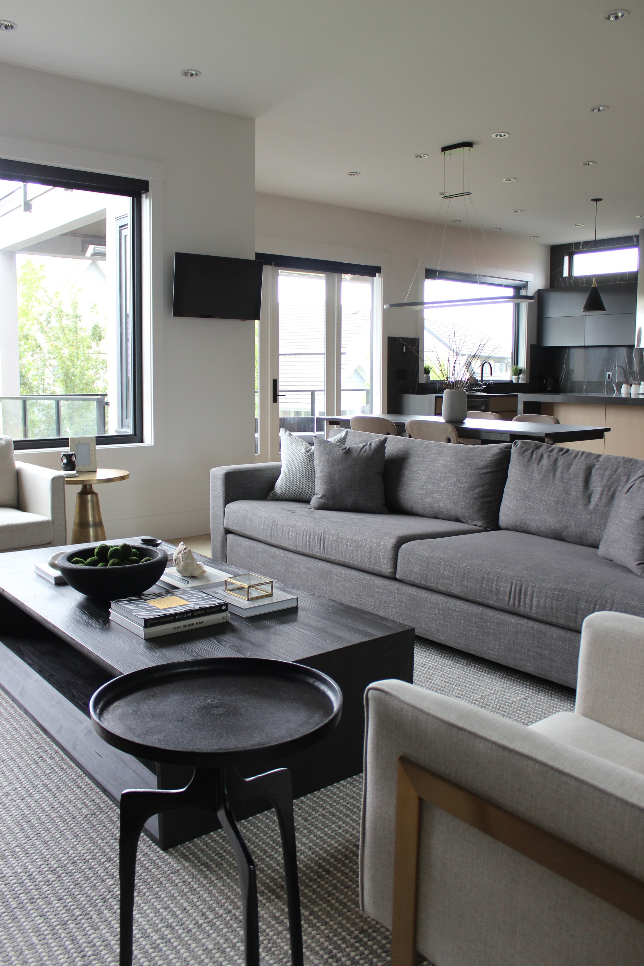 Modern living room with large windows, gray sofa, black coffee table, and a view of the kitchen in the background.