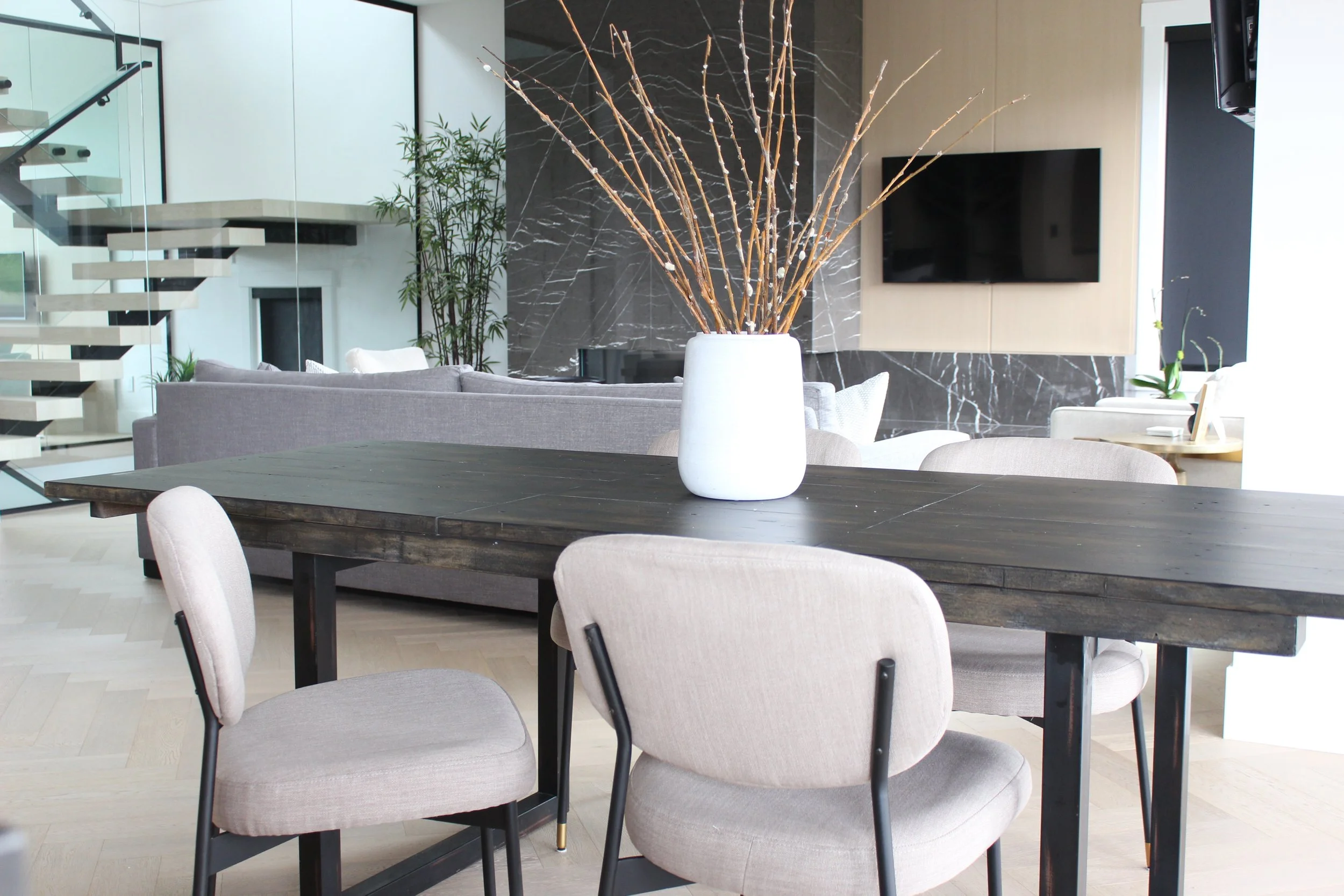 Modern living room with a dark wooden dining table, light upholstered chairs, a white vase with brown branches, a grey sofa, a black marble accent wall, a flat-screen TV, and a staircase with glass railing.