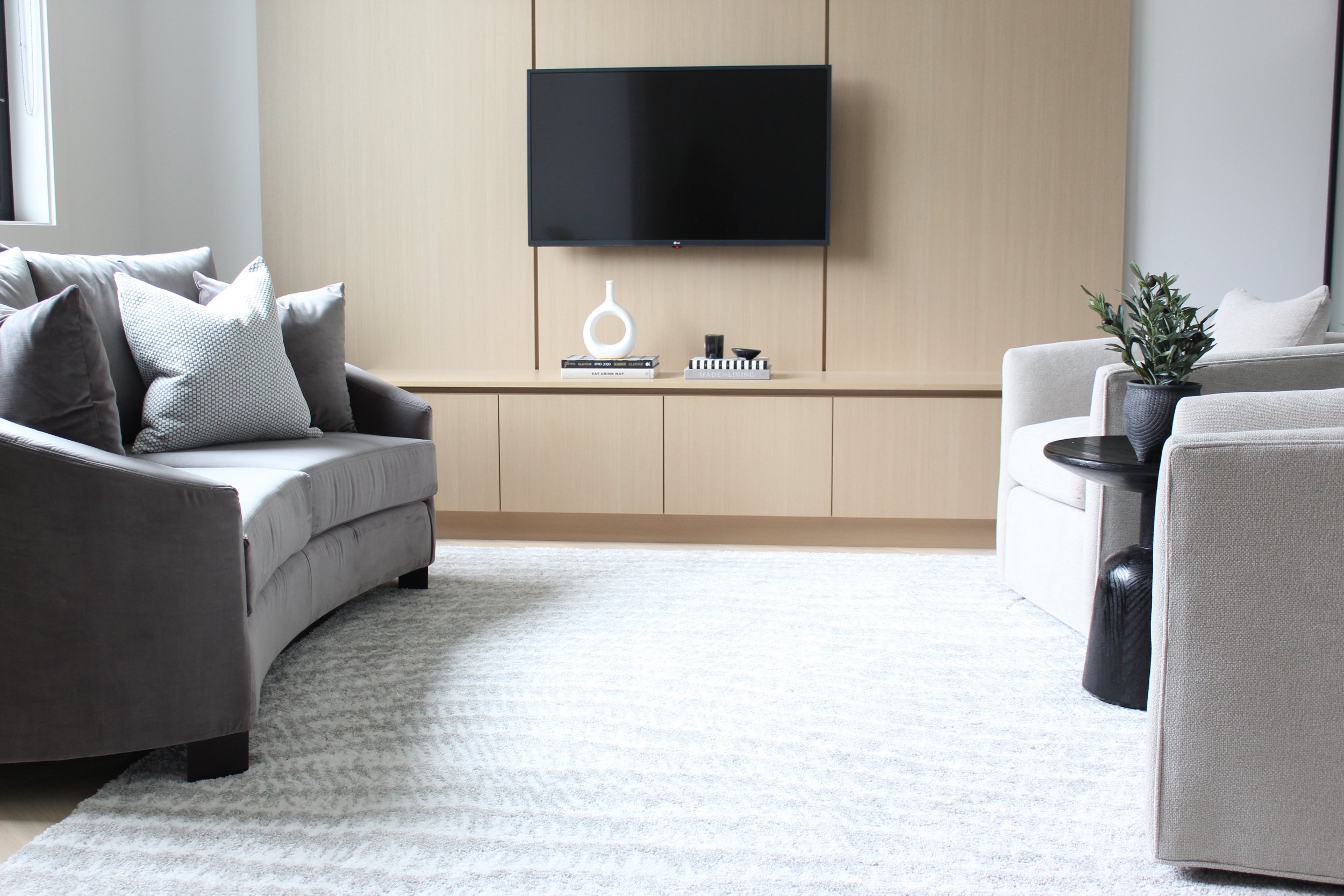 Modern living room with a gray sofa and pillows on the left, a white armchair with a potted plant on a side table on the right, a light wood entertainment unit with decorative items, and a wall-mounted black flat-screen TV on a beige wall.