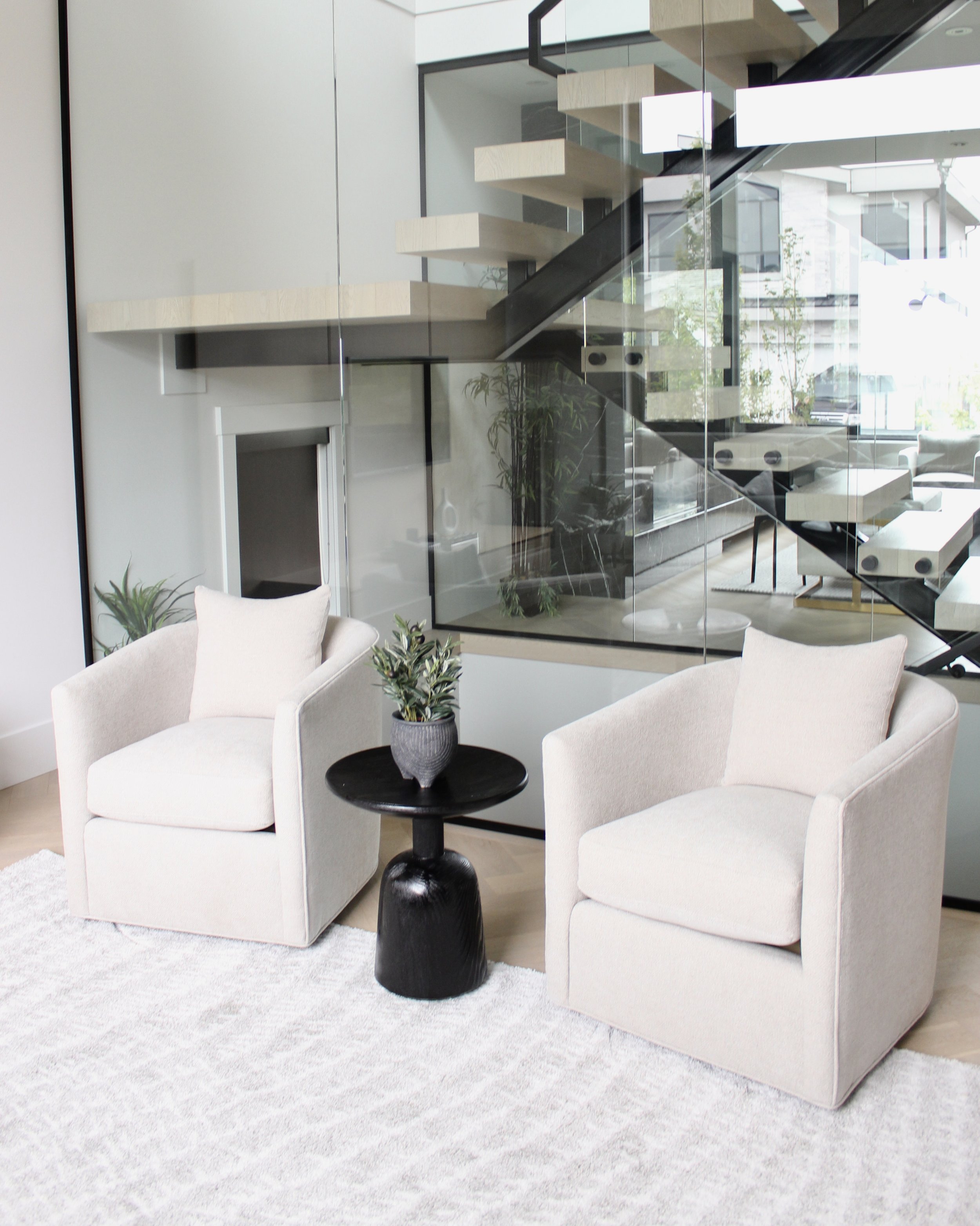 Modern living room with two beige armchairs, a small black side table with a potted plant, and a white textured rug. Behind, there's a glass wall with stairs and a reflection of the street outside.