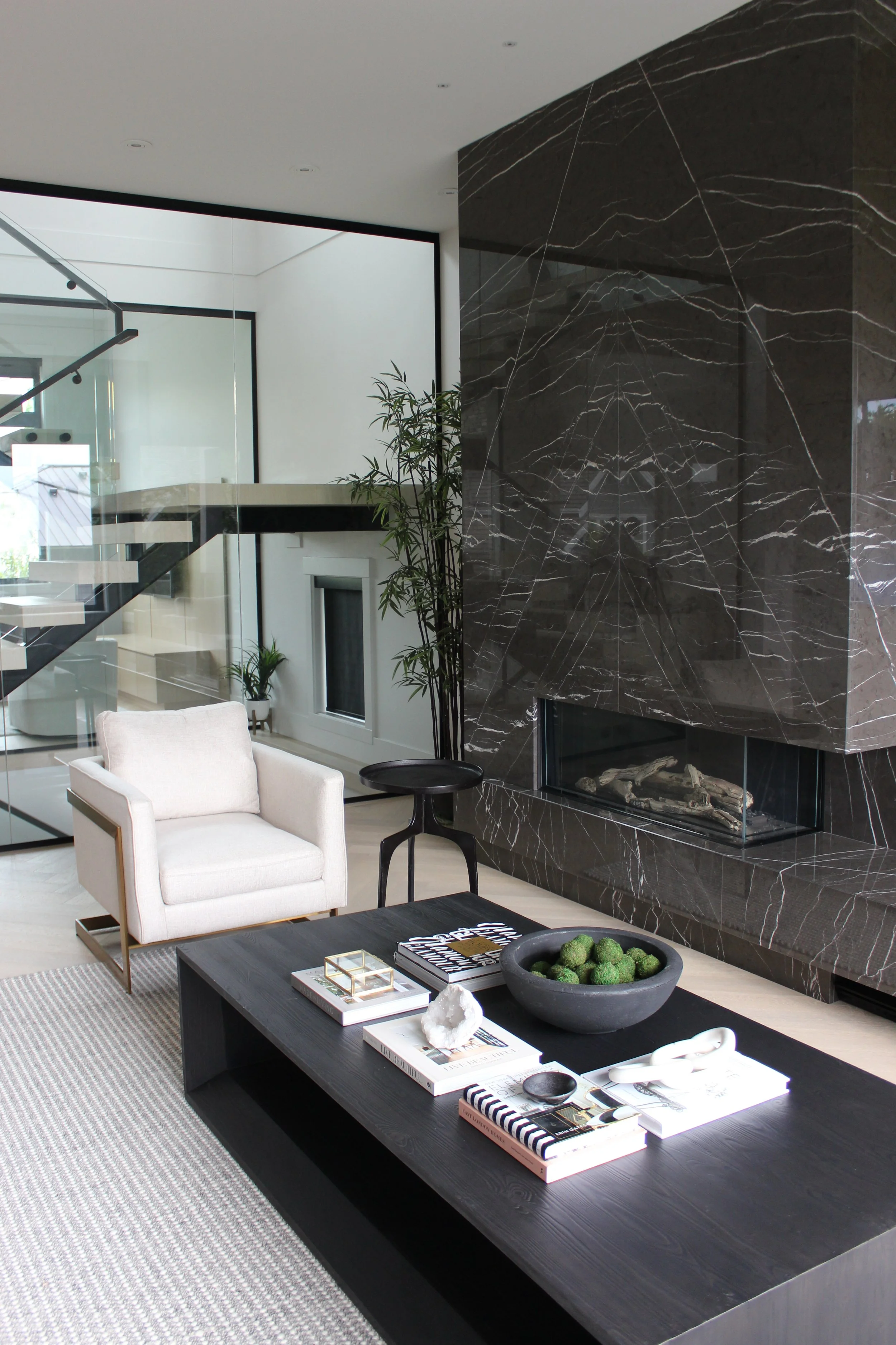 Modern living room with a black marble fireplace, white armchair, black coffee table, and staircase with glass railing.
