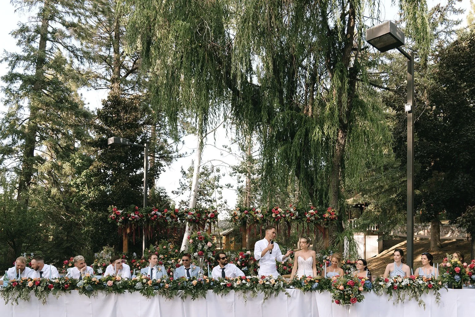 Bride and Groom stand at the Head table alongside the bridal party giving a thank you speech with a forest background and bright floral arrangements in Big Bear California