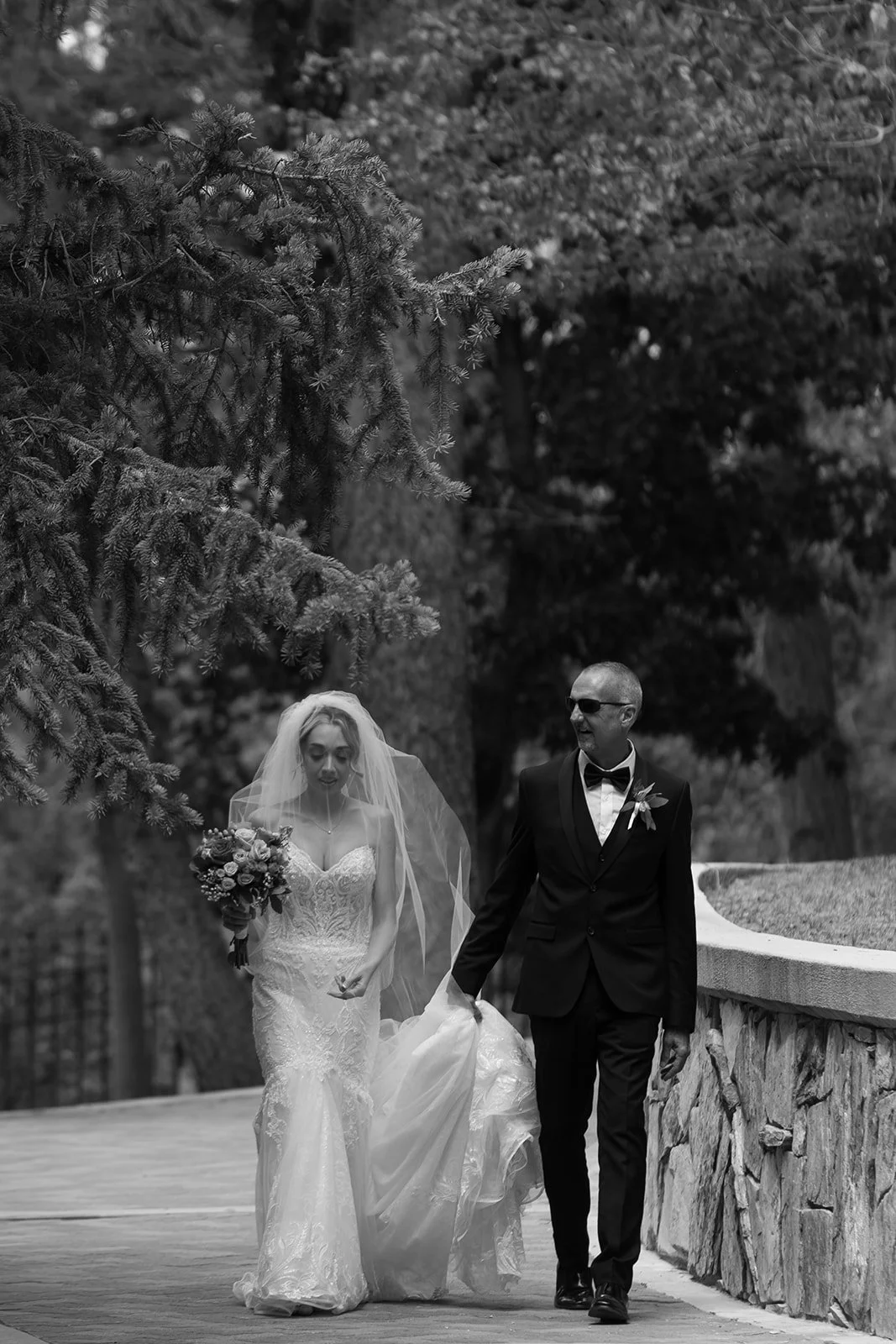Father of the Bride in a black tuxedo holding the bride's wedding dress train as they walk to the aisle together