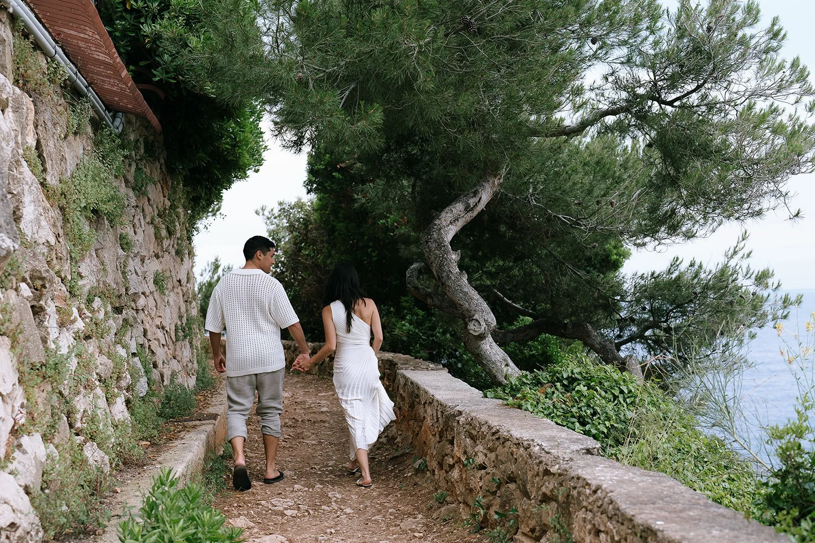 Couple holding hands walking along a stone trail surrounded by greenery and the Mediterranean Sea on the French Riviera