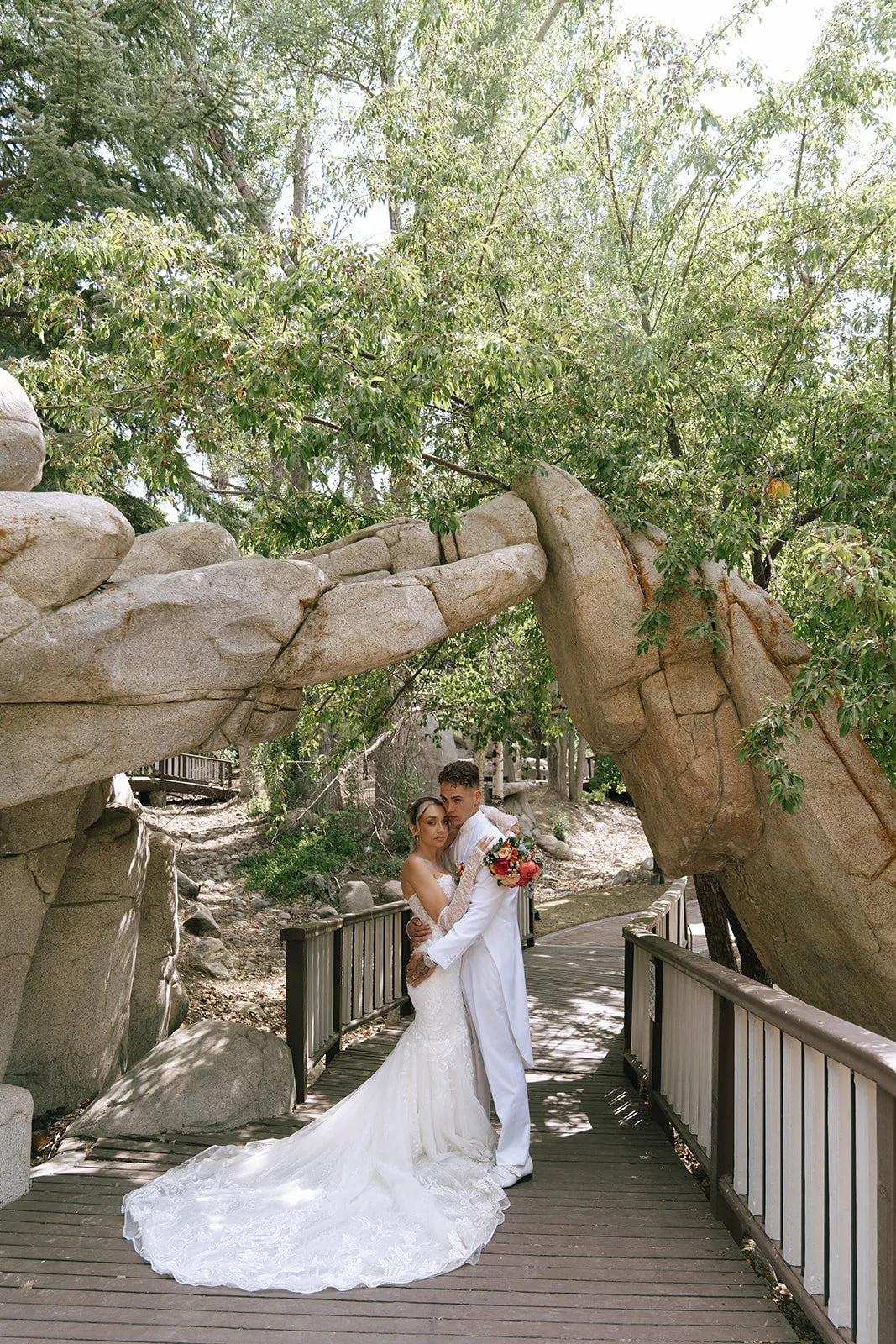 Bride and groom pose together on a bridge under a natural rock archway at Edgewood Mansion in Big Bear California, the bride holds a colorful bouquet.