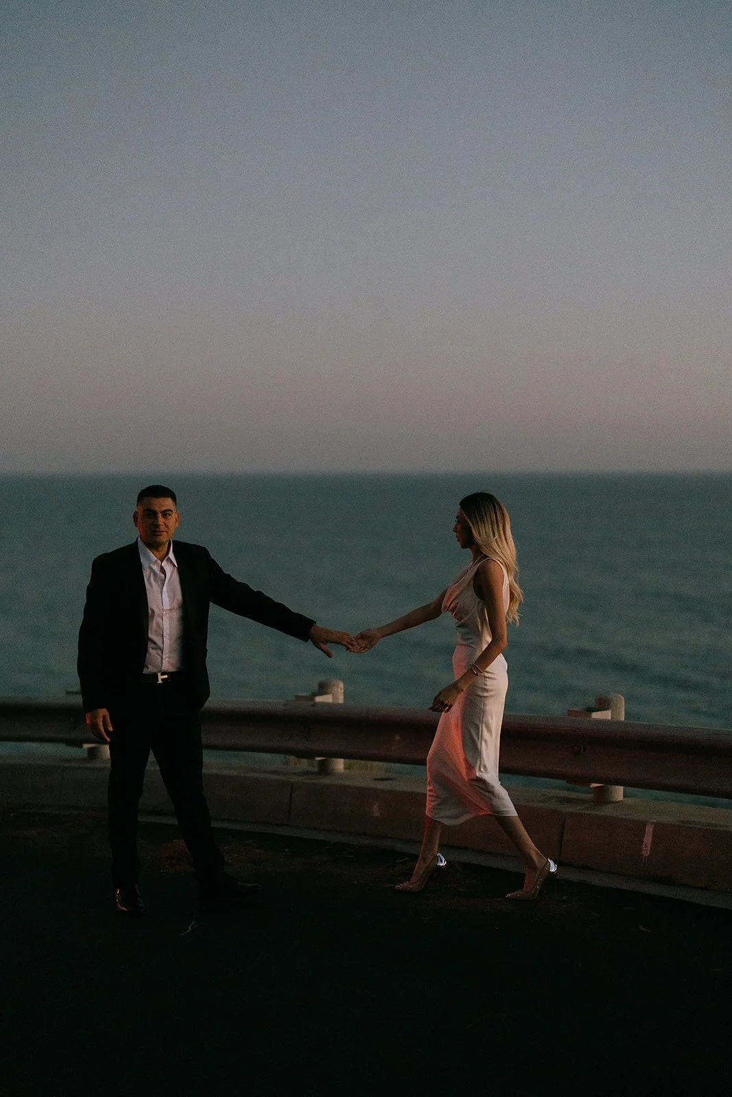 The couple holding hands with the ocean behind them