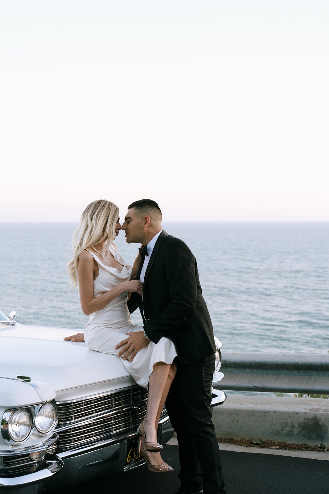 The bride wearing a silk gown and leaning on the car with the groom.