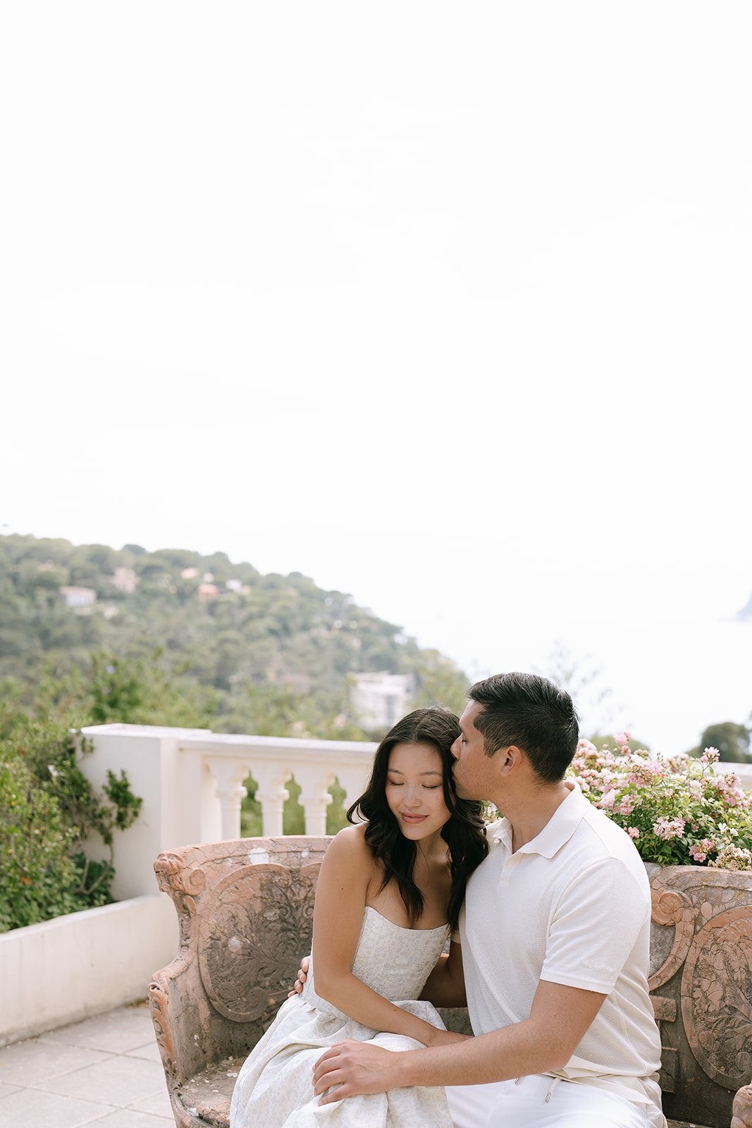 Groom to be kisses his bride to be on the forehead while sitting on a stone bench overlooking the Bay of Villefranche-sur-Mer