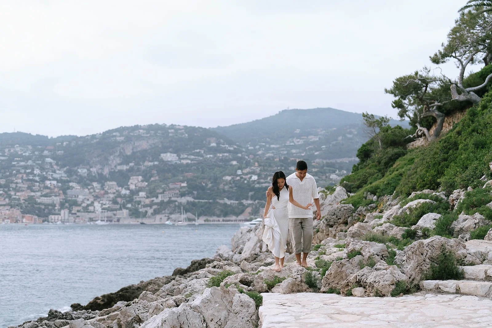 Couple walking along the coast of Saint-Jean-Cap-Ferrat with a view of Villefranche-sur-mer across the bay during their destination engagement session