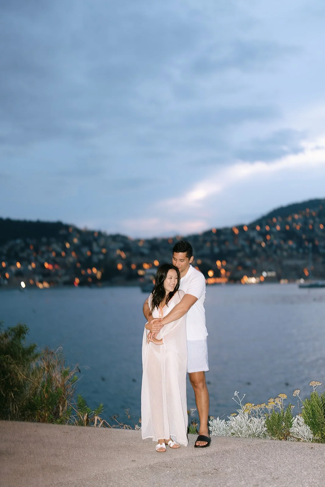 Couple standing together with the lights of the villas of the Villefranche-sur-Mer shining behind them during their coastal destination engagement photoshoot on the South of France