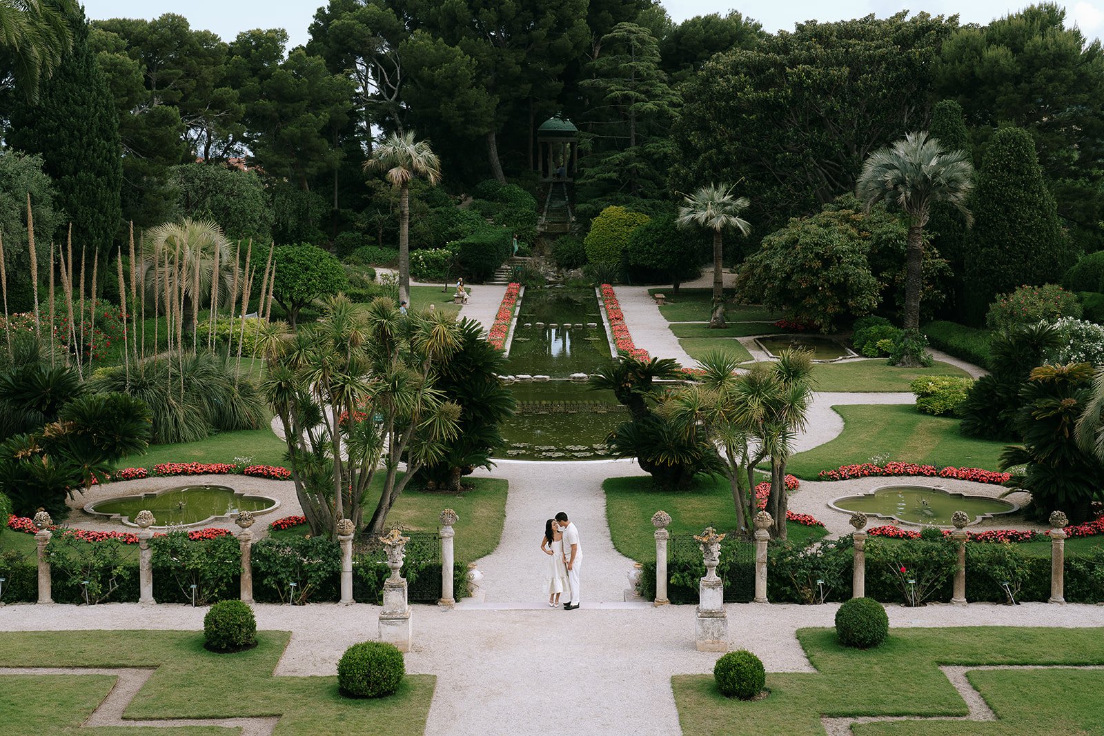 Wide shot of couple kissing in the French garden surrounded by pink flower beds, lush greenery, and statues in the Renaissance style of the Villa Ephrussi