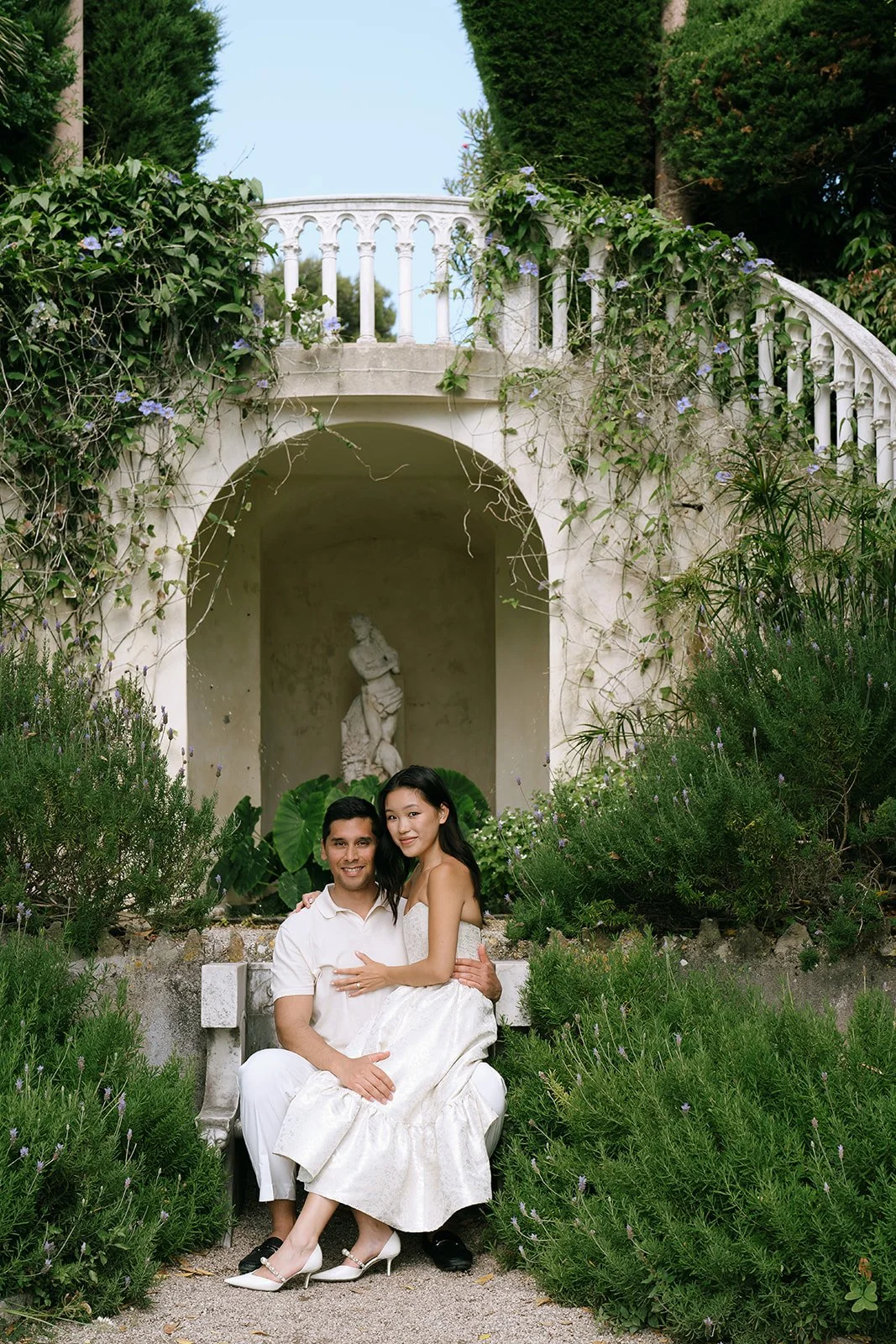 Soon to be bride and groom sit together on a stone bench surrounded by a statue and a horseshoe staircase in the Florentine garden of the Villa Ephrussi