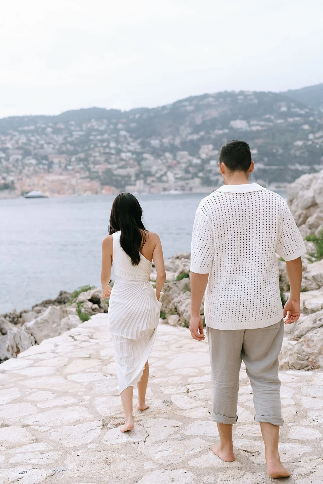 Couple walking barefoot on a cobblestone path towards the Bay of Villefranche during their destination engagement session on the Saint-Jean-Cap-Ferrat coastline
