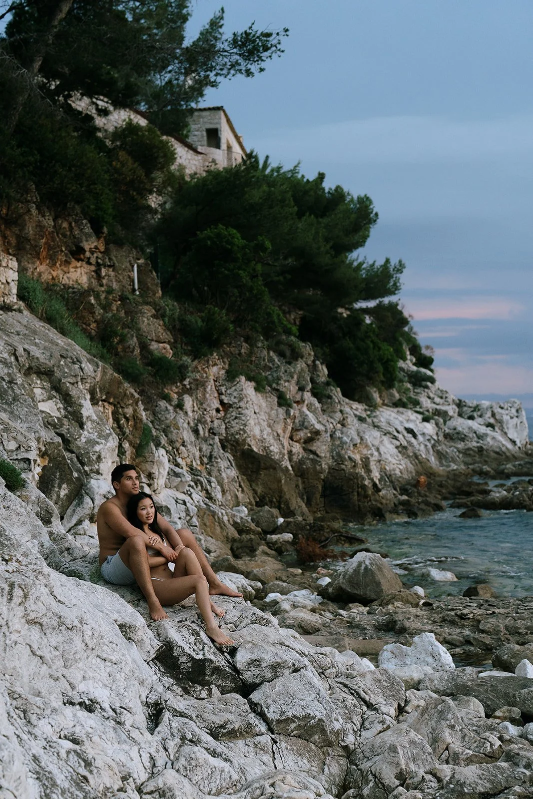 Couple in white swimsuits sitting on rocks of the Plage de Passable watching the sunset on the Mediterranean coast during their destination engagement photoshoot