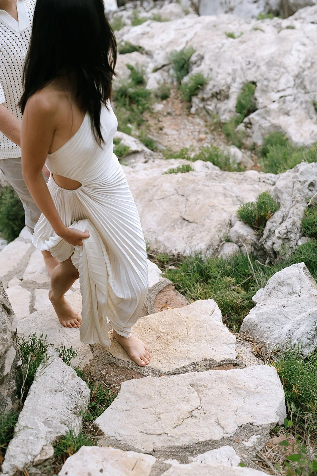 Soon to be bride holds her white pleated dress while she walks up a stone staircase along the Mediterranean coast in the South of France