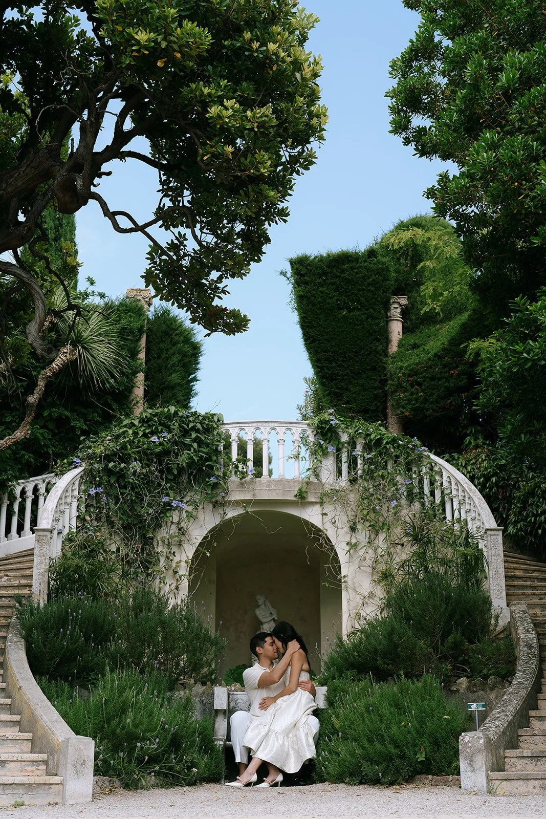 Soon to be bride and groom sit together on a stone bench surrounded by a statue and a horseshoe staircase in the Florentine garden of the Villa Ephrussi