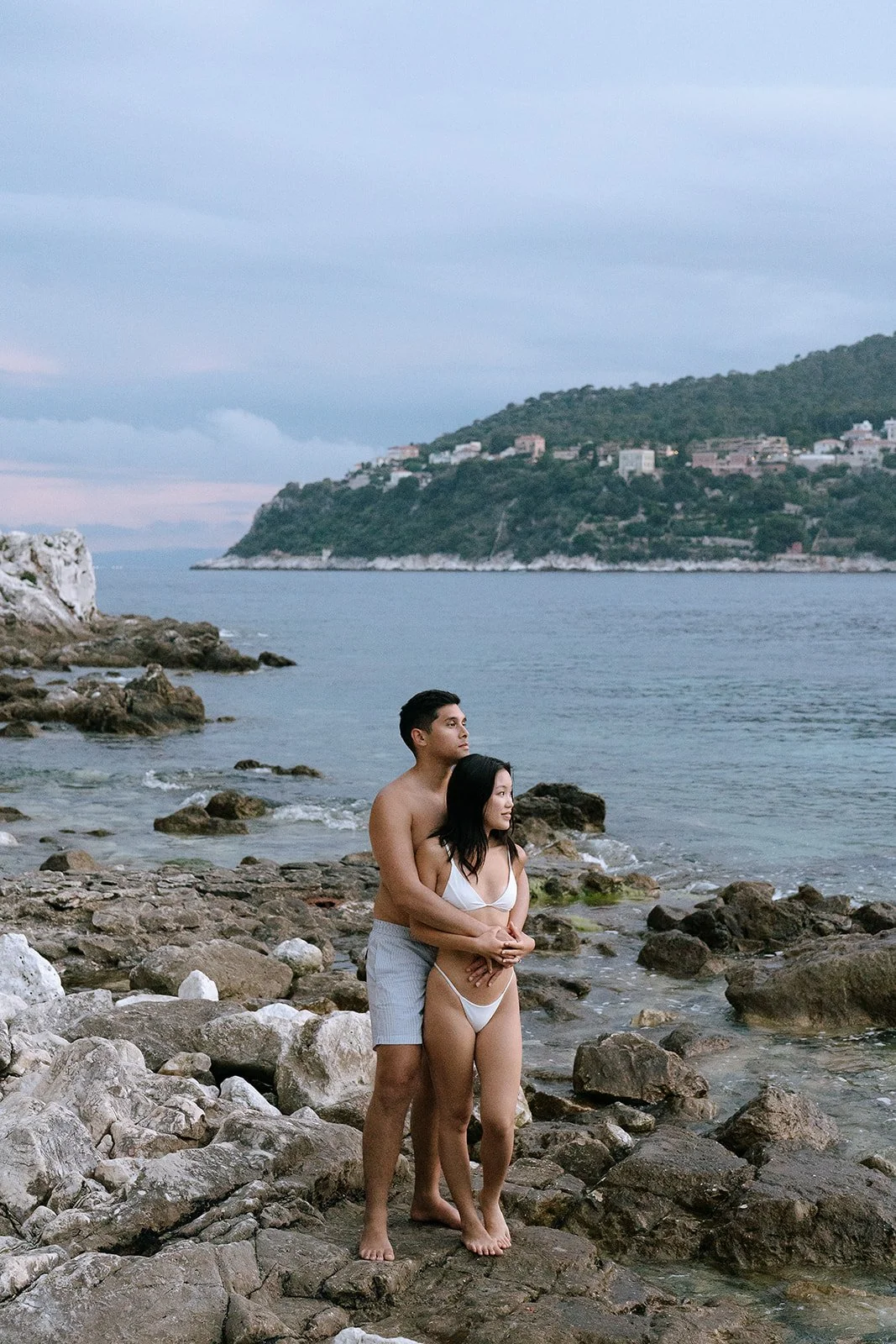 Couple in white swimsuits standing together on Passable Beach watching the sunset over the Bay of Villefranche during their destination engagement photoshoot on the South of France