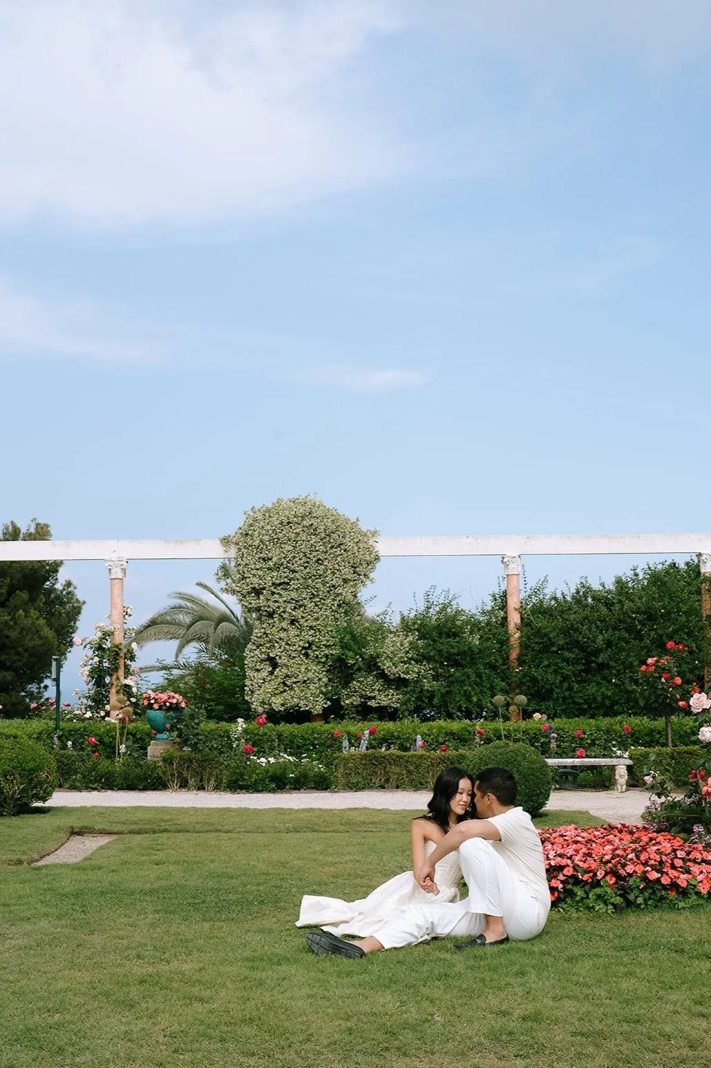 Couple sits together on the grass surrounded by pink flower beds in the French Garden of the Villa Ephrussi for their destination engagement photoshoot