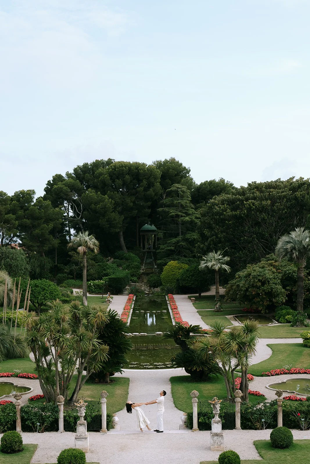 Wide shot of couple kissing in the French garden surrounded by pink flower beds, lush greenery, and statues in the Renaissance style of the Villa Ephrussi