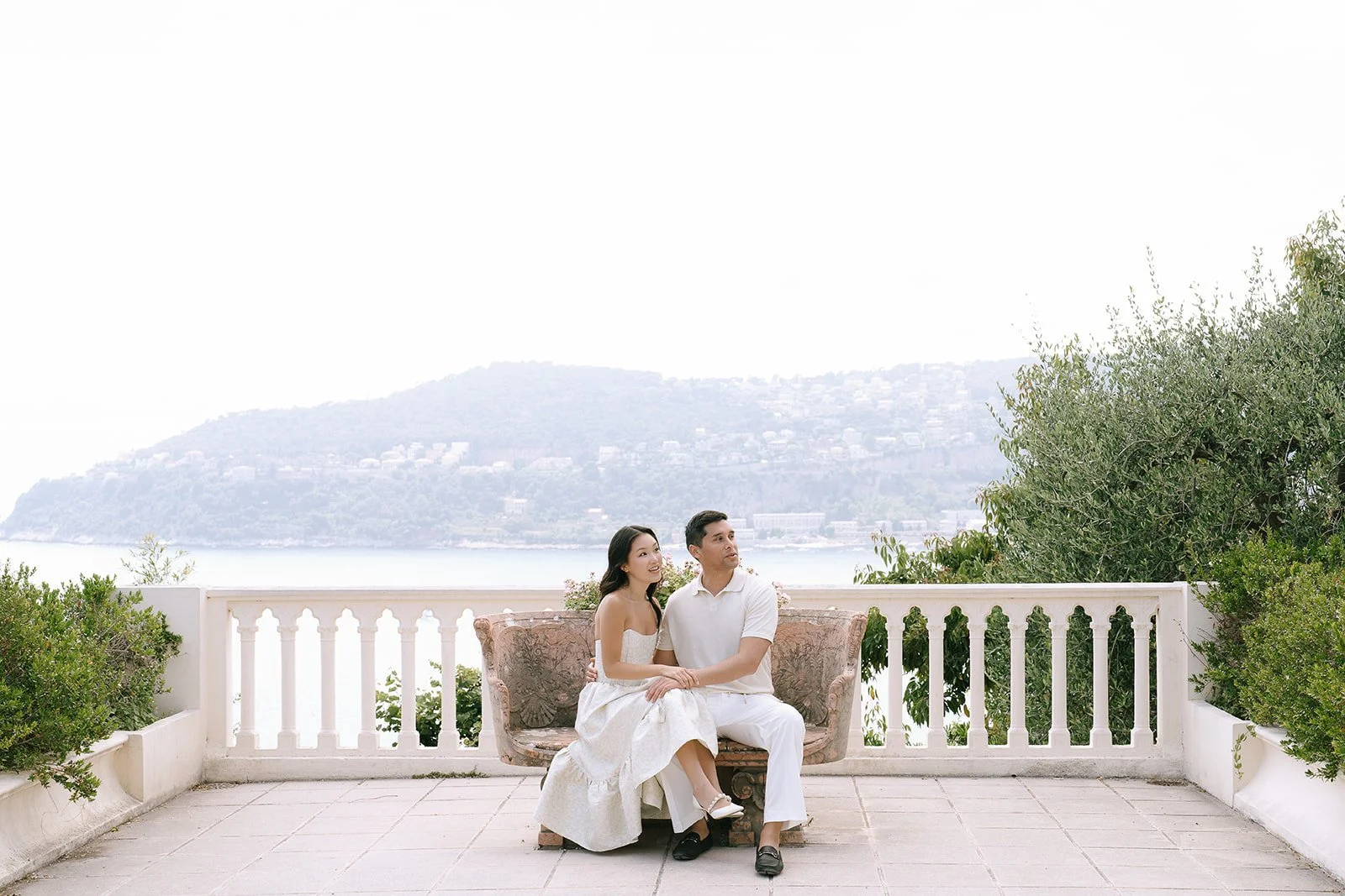 Soon to be bride and groom sit together on a stone bench on a balcony overlooking the Rade de Villefranche Sur Mer or the Bay of Villefranche-ser-Mer on the Mediterranean coast