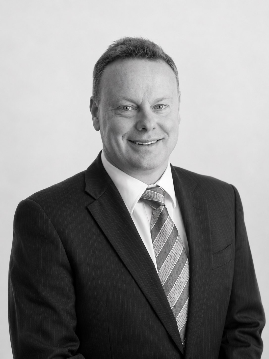 Black and white professional headshot of a man in a suit and tie, smiling, against a plain background.