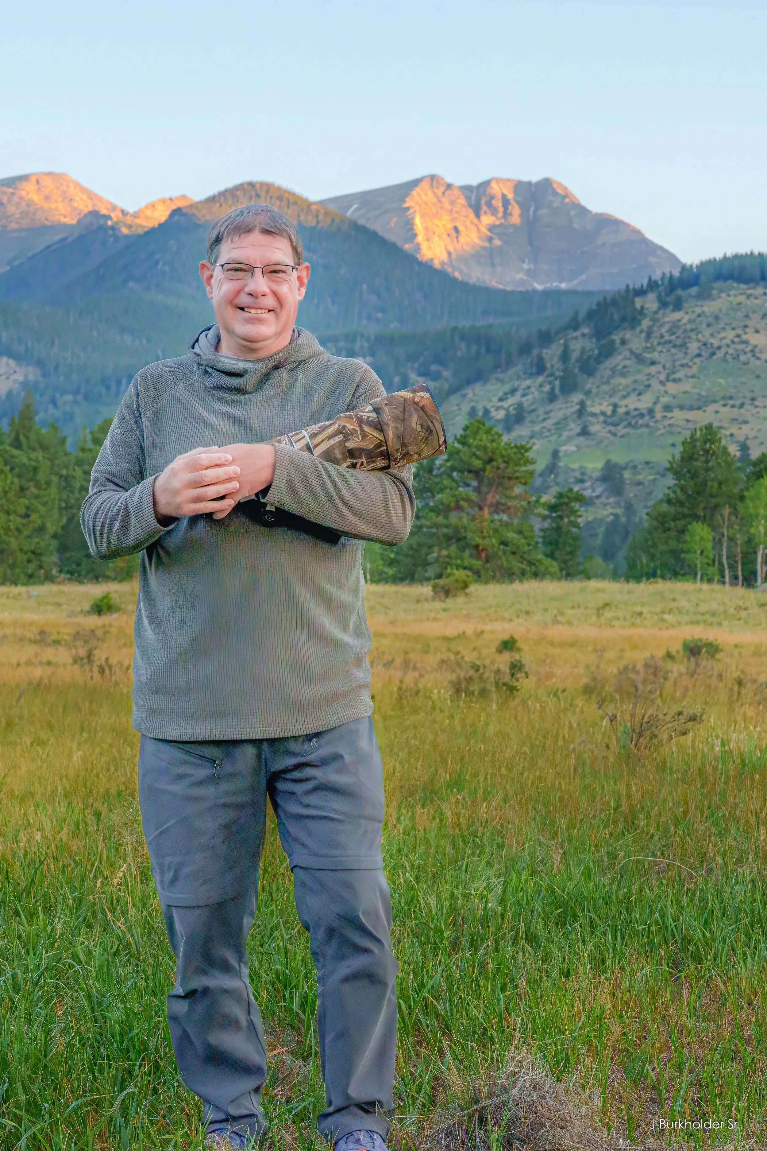 A man standing in a grassy field with mountains in the background during sunset, smiling, wearing glasses and a long-sleeve shirt, with his arms crossed.
