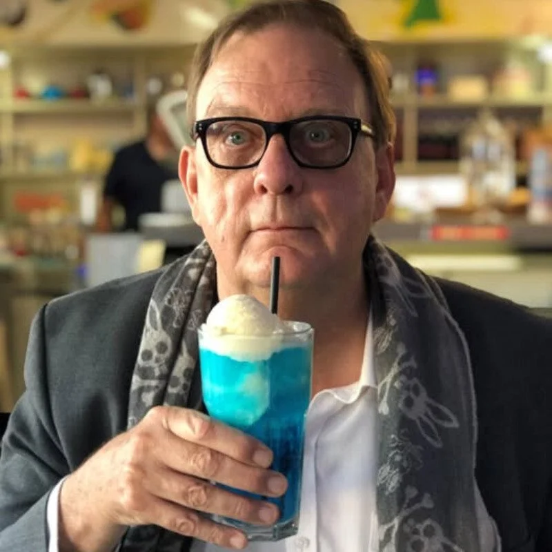 Peter Goers wearing black rimmed glasses looking at the camera with a bright blue drink topped with vanilla icecream and a straw in his right hand lifting towards his mouth