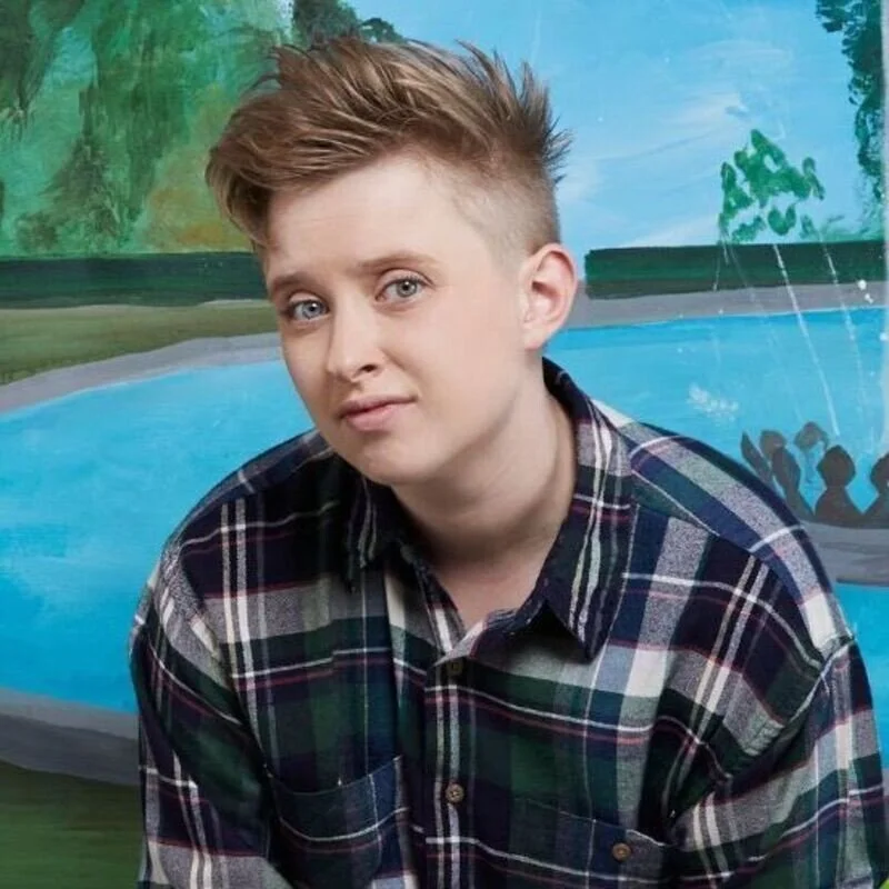 Hanna in a blue, white and red checked button down shirt in front of a painted background depicting a river scene