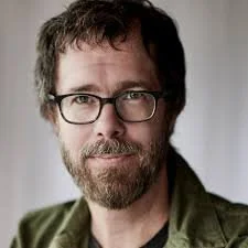 Headshot of Ben Folds with short brown hair, glasses and a beard, wearing a green jacket over a dark shirt, looking at the camera against a softly lit background.