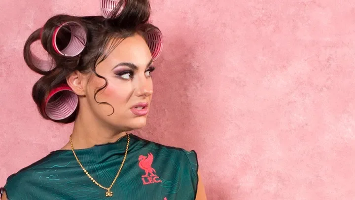 Jade Franks, curlers in hair, green top with gold chain around her neck. Pink mottled background.