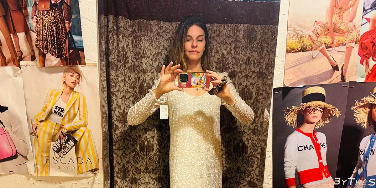 Woman taking a mirror selfie in a fitting room, wearing a cream lace dress, surrounded by fashion posters on the walls and patterned fabric behind her.
