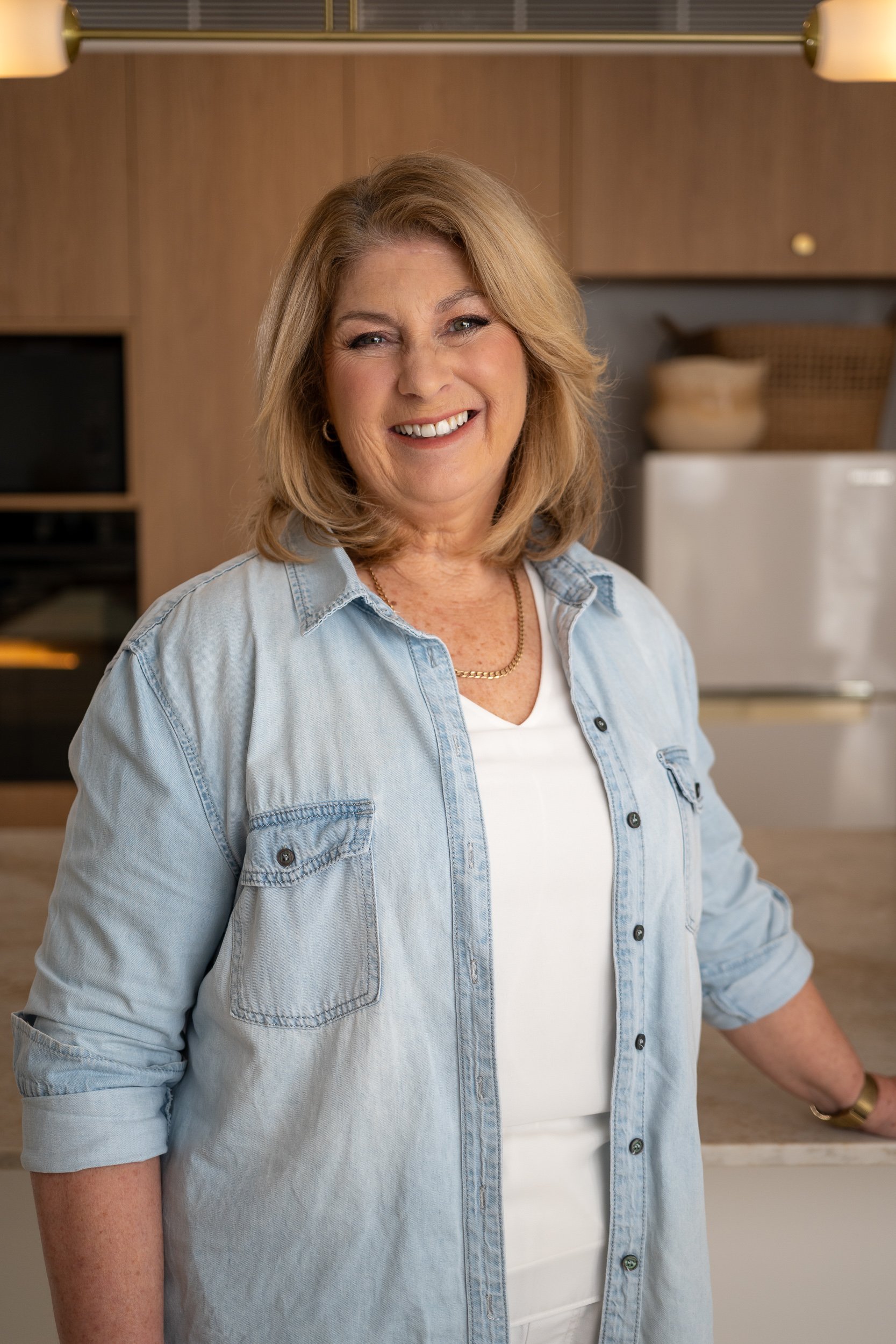 Smiling woman with blonde hair wearing a light denim shirt over a white top, standing in a modern kitchen.
