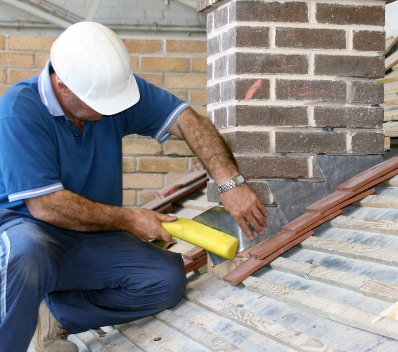 A construction worker installing roof tiles on a house roof with a brick chimney nearby.