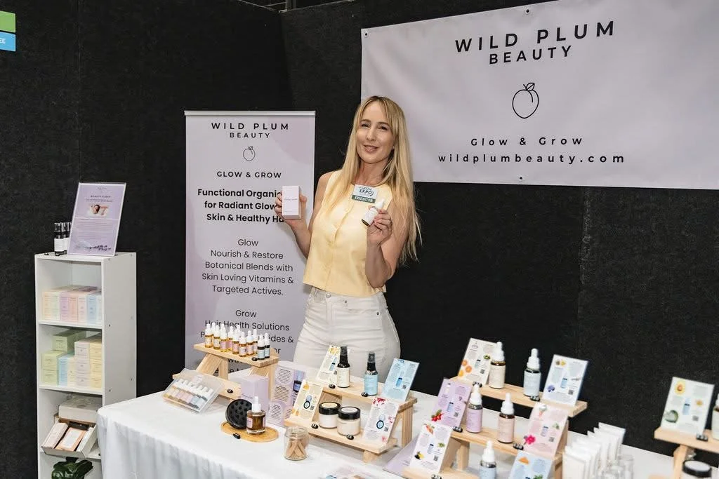 A woman at a trade show booth for Wild Plum Beauty, holding a product and smiling. The booth displays skincare items and promotional materials.