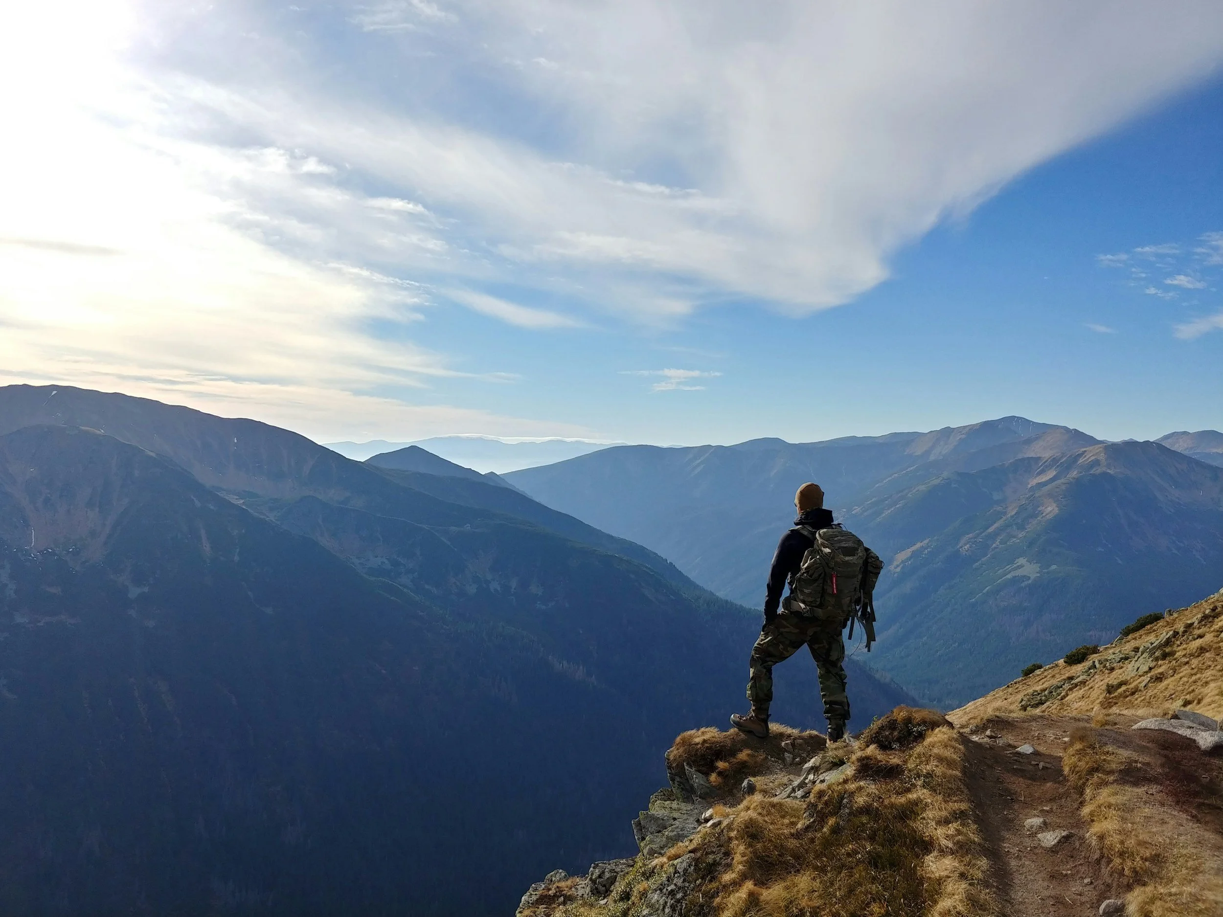 A hiker with a backpack standing on a mountain ledge, looking across a range of mountains under a partly cloudy sky.