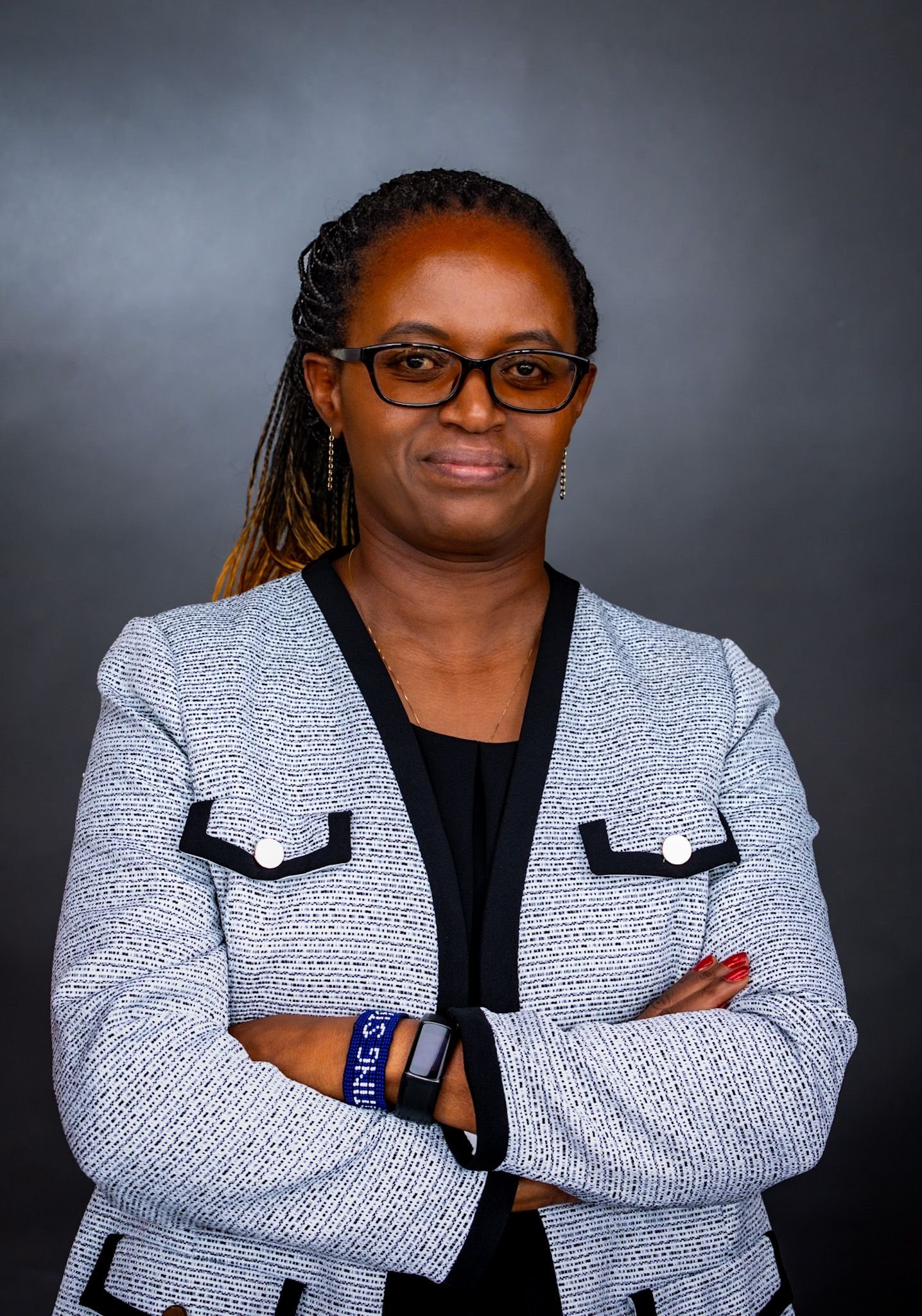 A woman with glasses and braids, crossing her arms and smiling, wearing a gray and black patterned blazer and a smartwatch against a dark background.